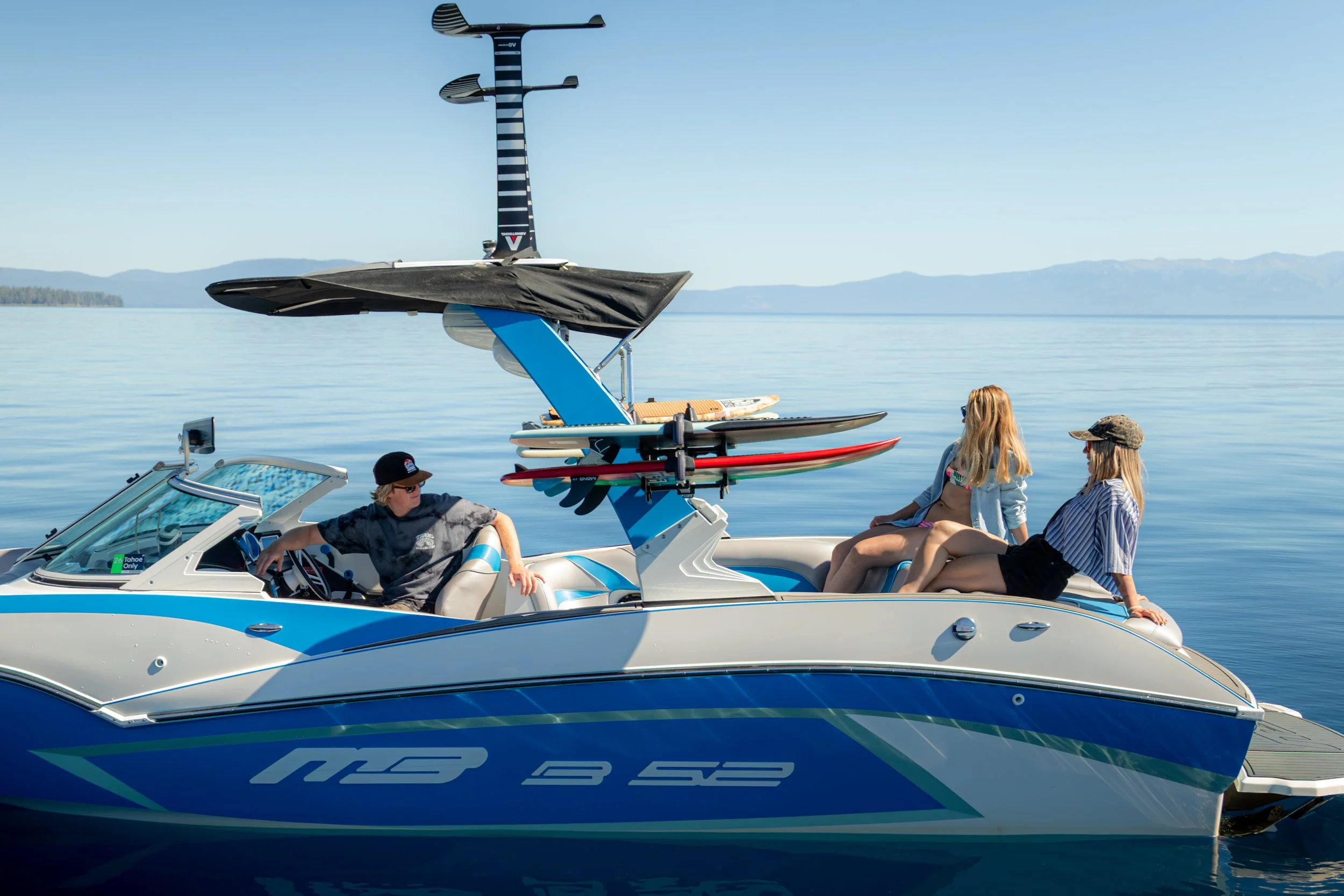 A group of people on a luxury motorboat on a calm lake, with a wakeboard boat in the background carrying several wakeboards, and a scenic mountainous landscape in the distance.