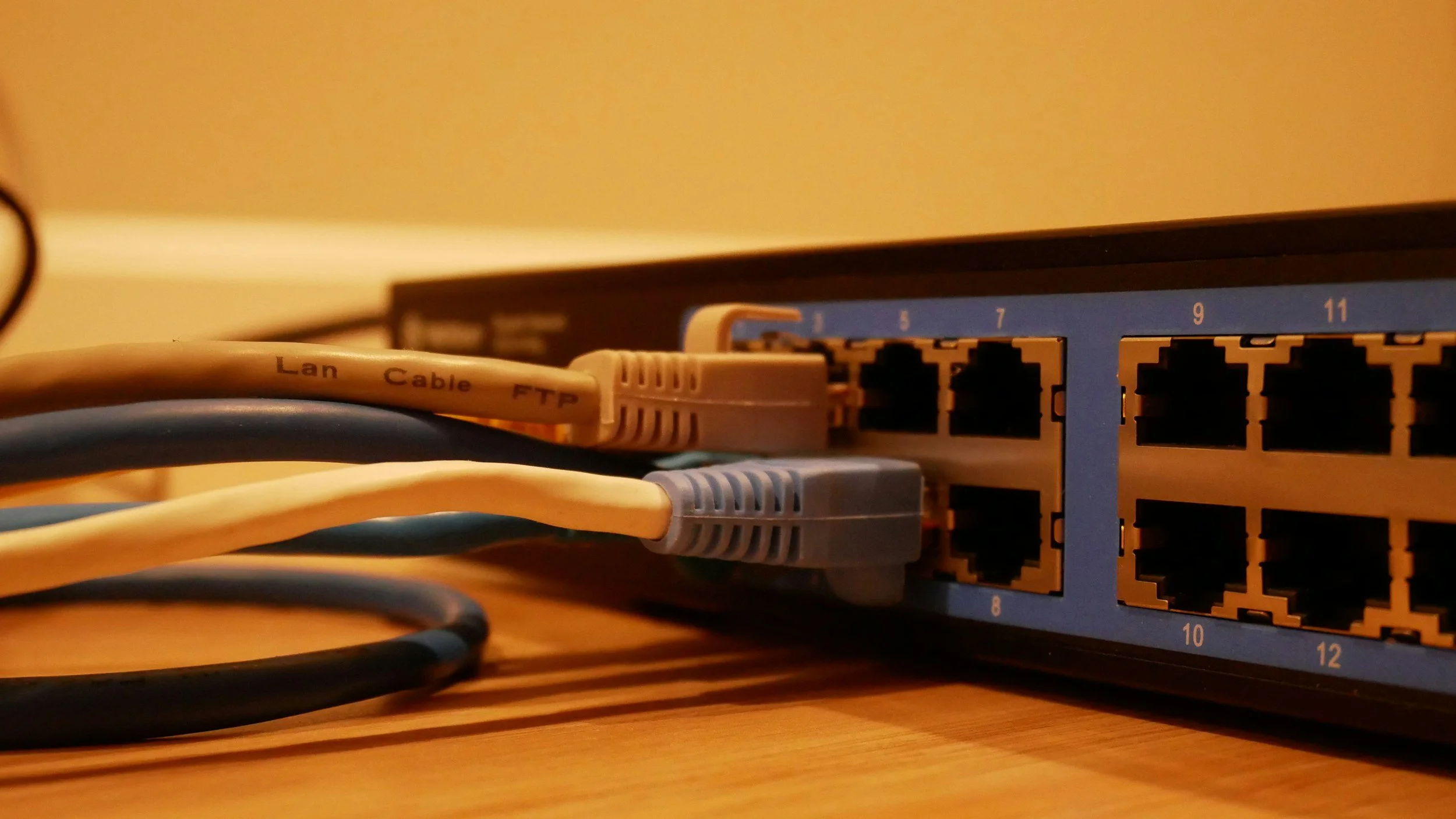 Close-up photo of a network switch with several Ethernet cables plugged into it. The cables are labeled 'Lan,' 'Cable,' and 'FTP.' The switch is on a wooden surface, with a blurred background.