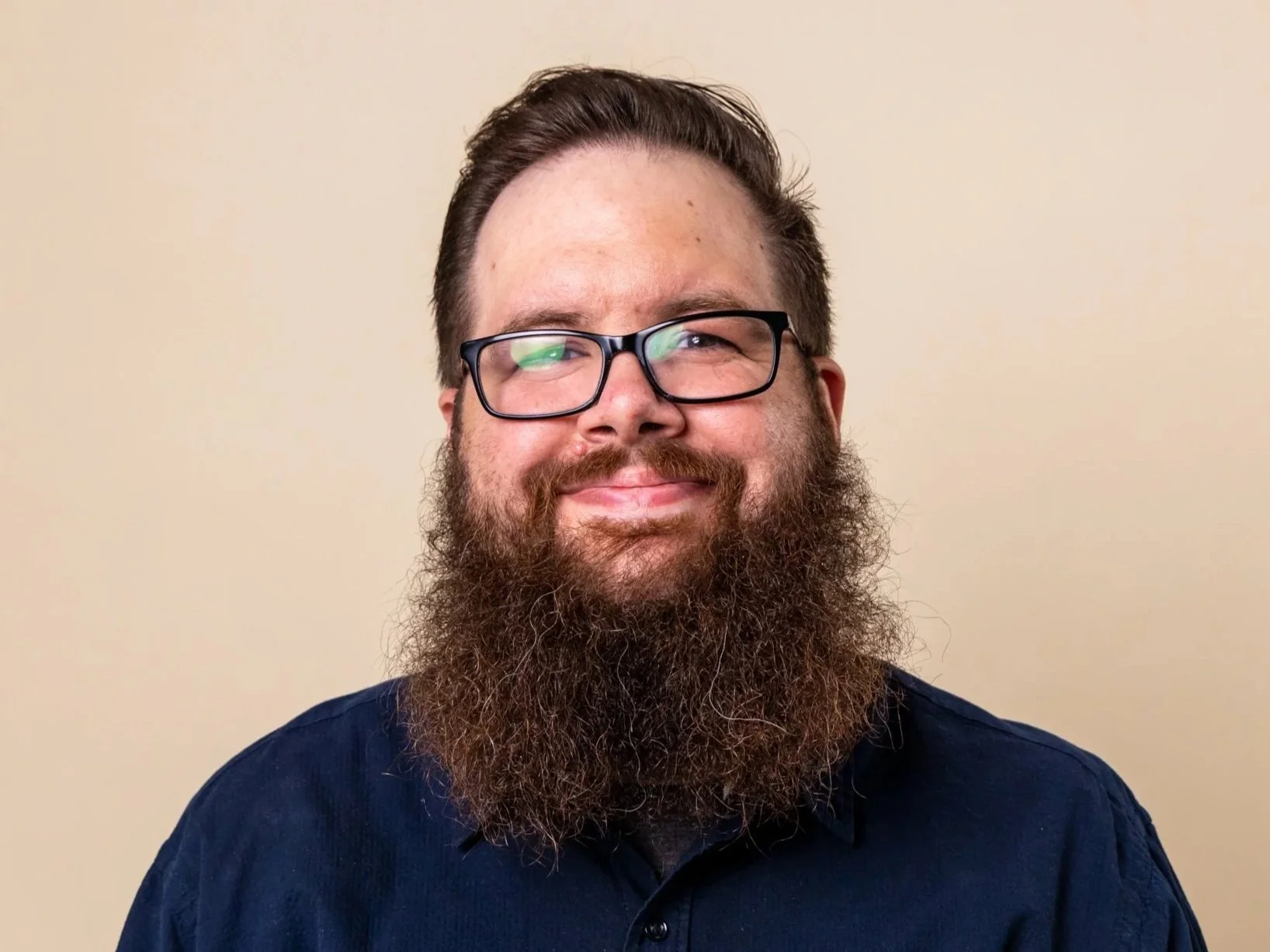 A man with glasses and a long beard, smiling against a beige background.