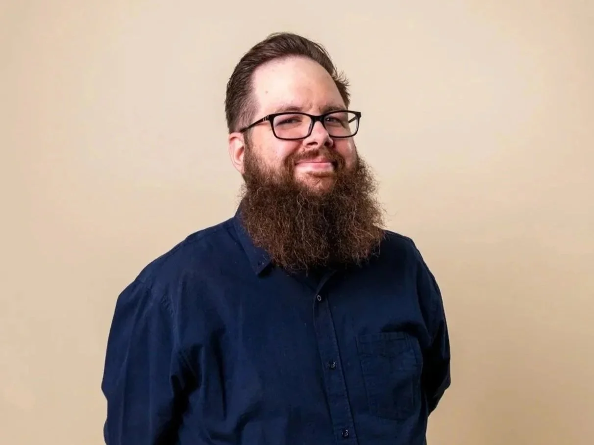 A man with glasses, a beard, and brown hair wearing a dark blue shirt against a plain beige wall.