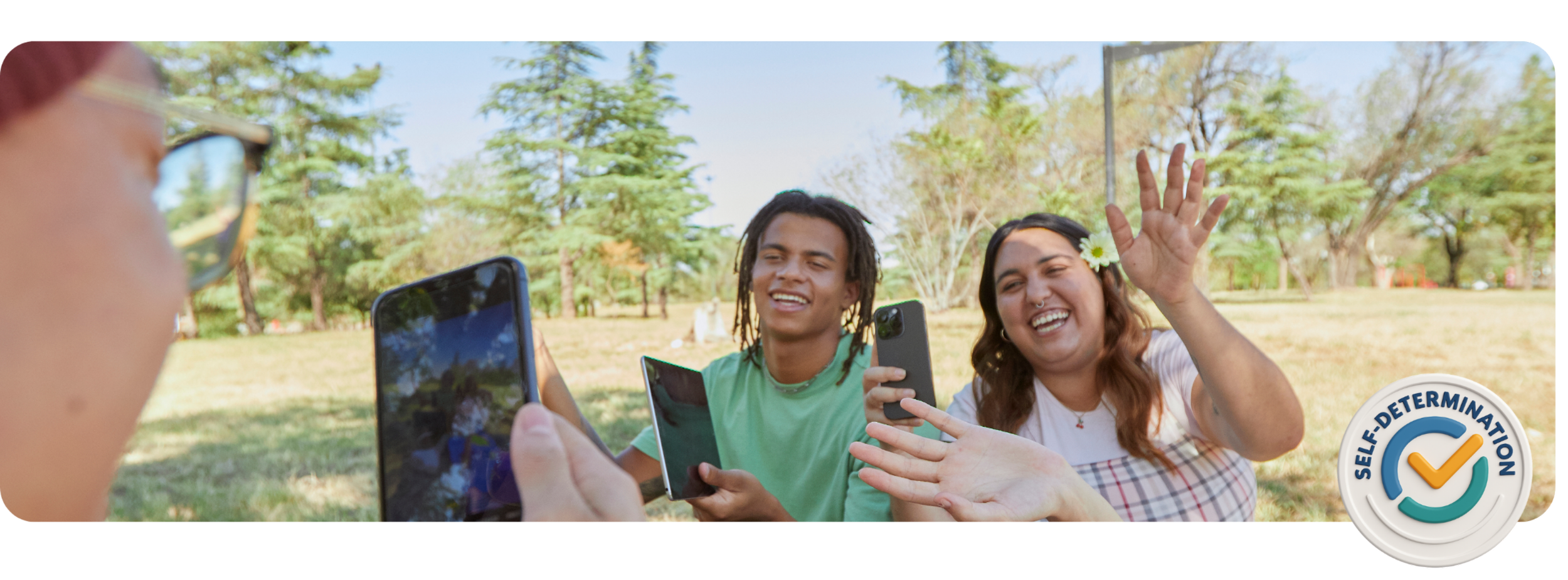 Tres amigos al aire libre tomándose selfies y sonriendo, una mujer saludando, con una insignia que dice "Autodeterminación" en la parte inferior derecha.