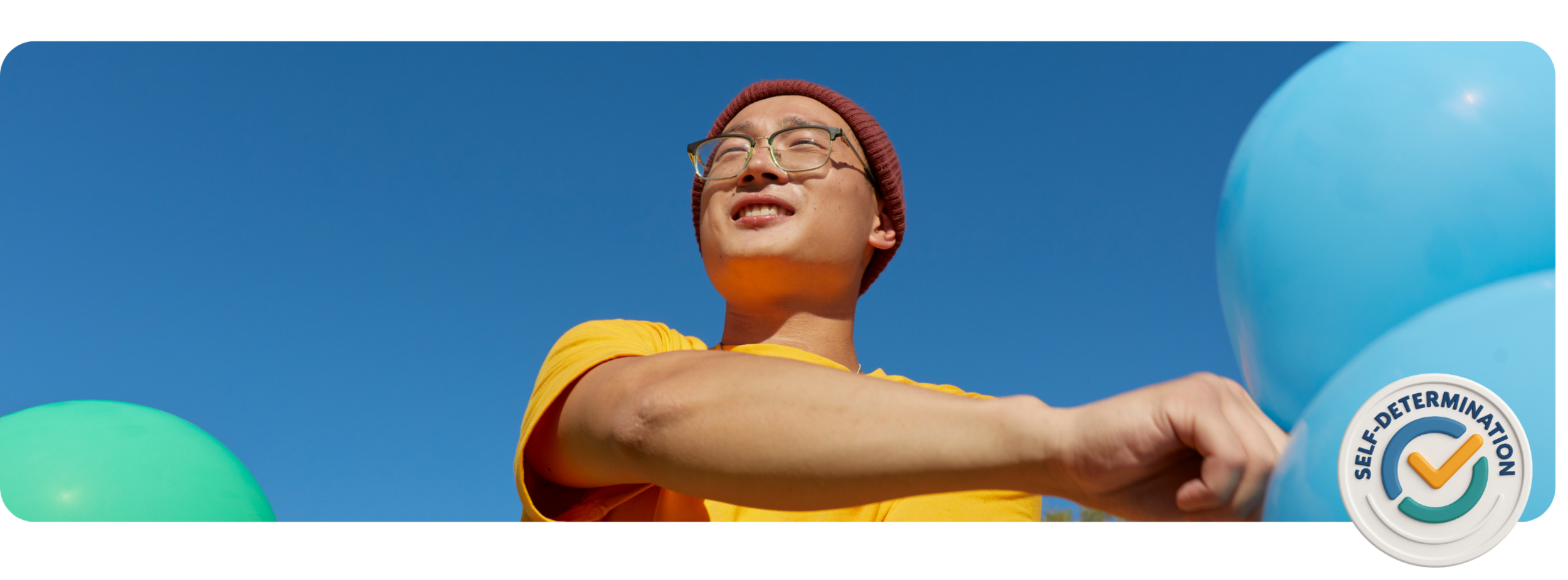 Young man with glasses and a maroon beanie smiling outdoors against a clear blue sky, holding blue and green balloons, with a 'Self-Determination' badge on his yellow t-shirt.