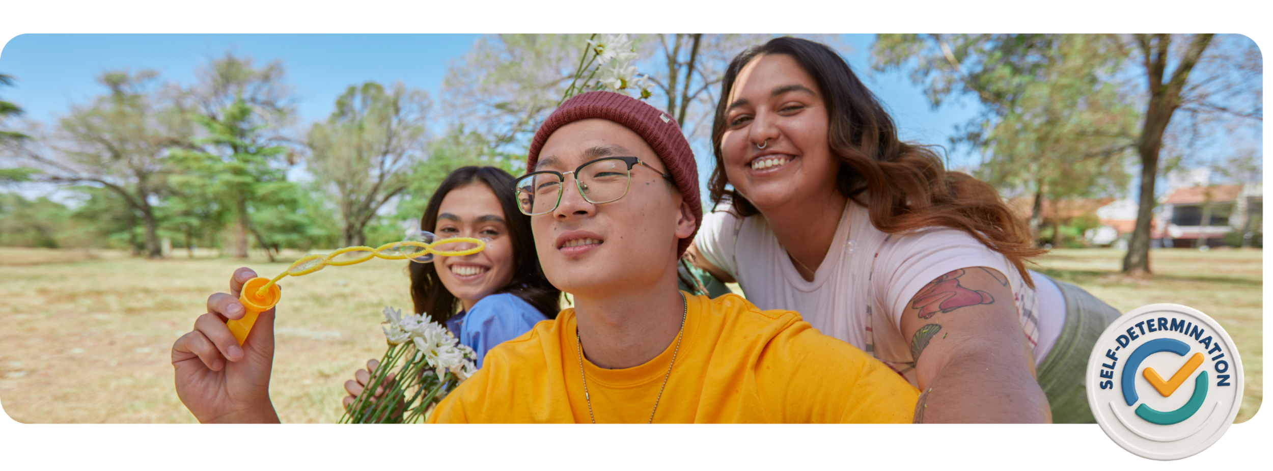 Group of three friends taking a selfie outdoors in a park, with trees and blue sky in the background, smiling and holding flowers and bubble-blowing toys.