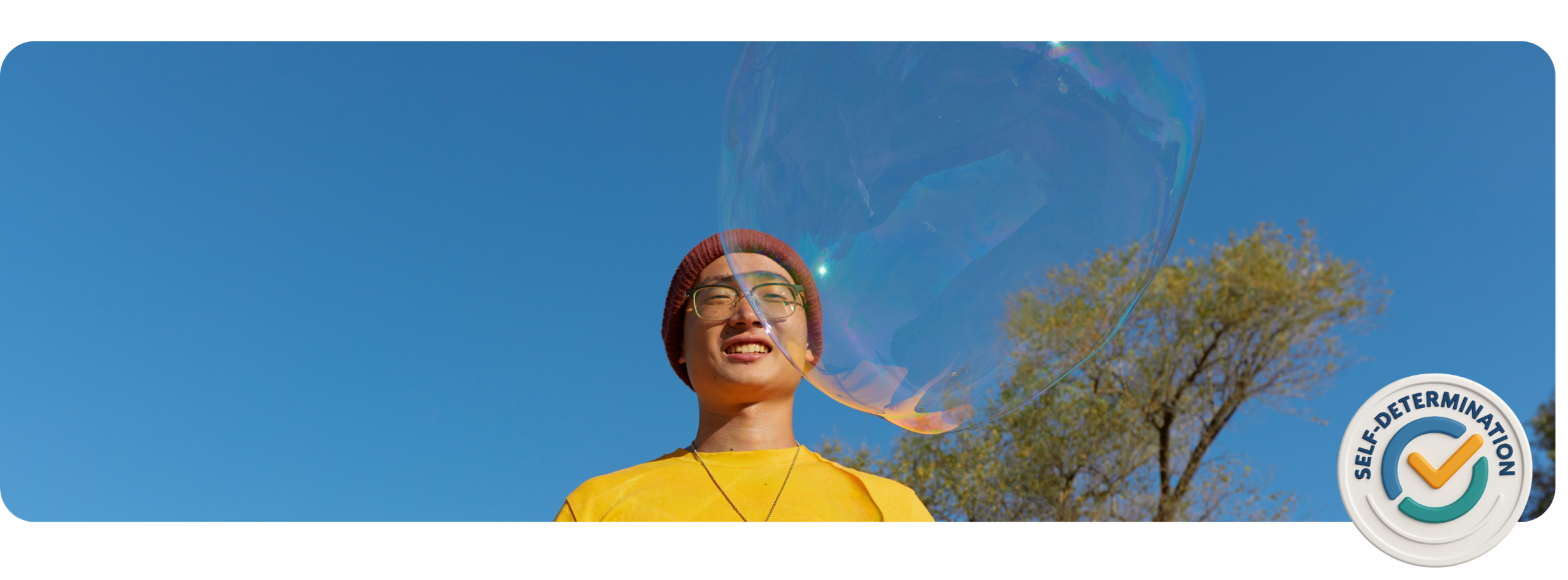 A smiling person with glasses and wearing a red knit hat and yellow shirt, looking at a large soap bubble outdoors with blue sky and trees in the background.