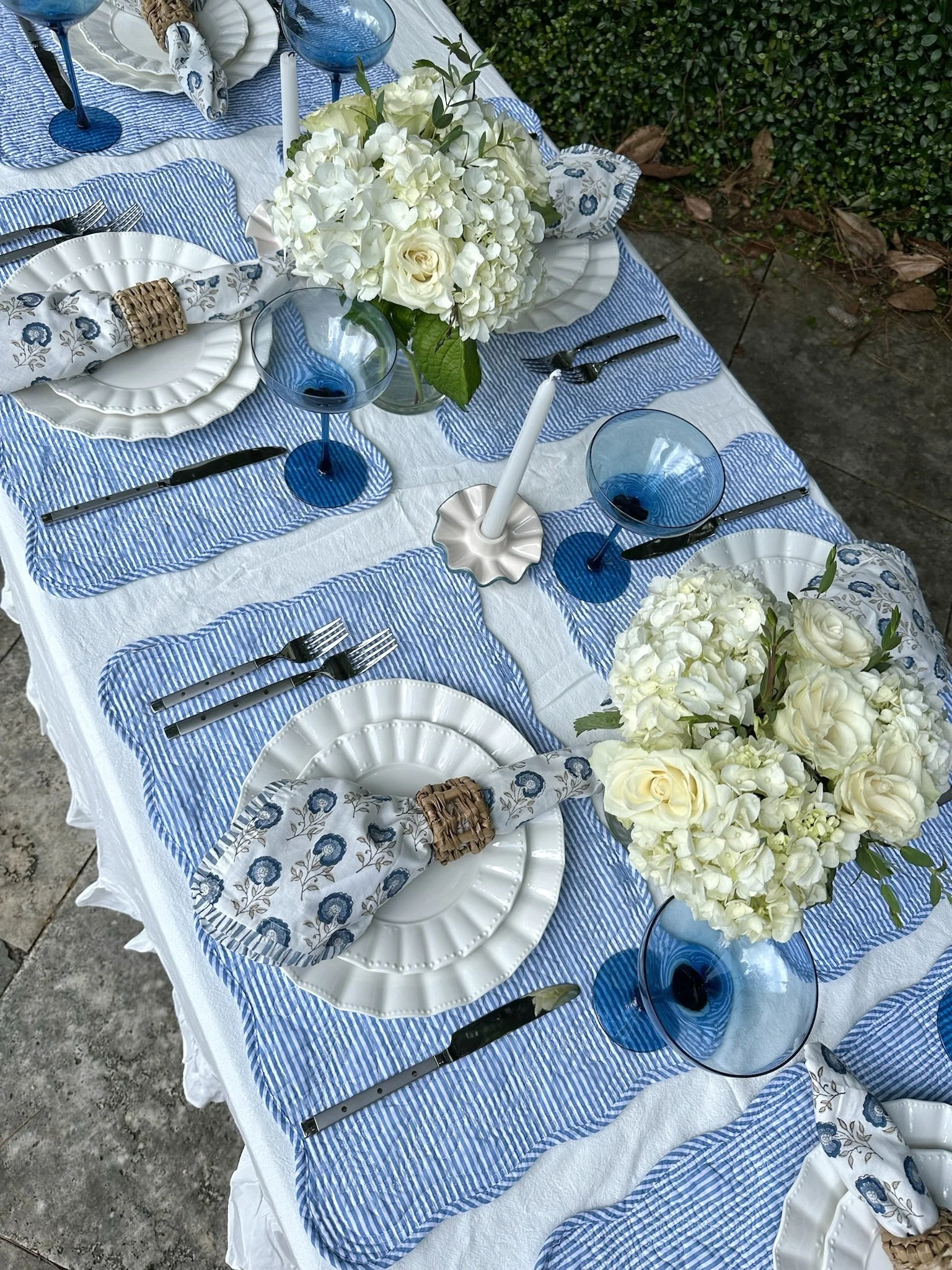 A table set for a meal with white plates, silverware, blue wine glasses, white floral napkins in patterned rings, white floral centerpieces, and blue striped placemats.