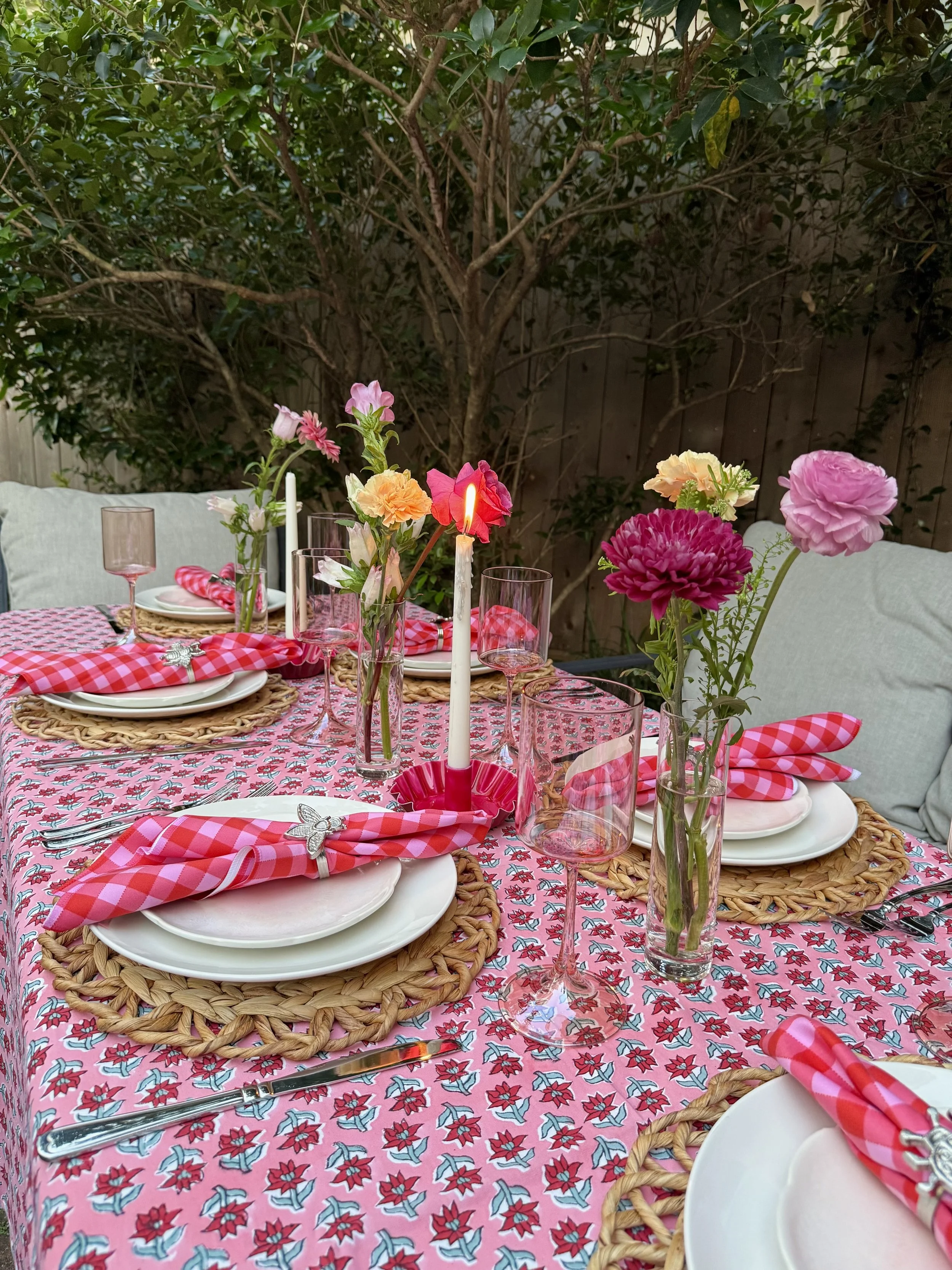 A beautifully set outdoor table with pink floral patterned tablecloth, white plates on woven placemats, pink checking napkins with silver napkin rings, pink wine glasses, and a tall white candle in the center surrounded by small vases with pink and peach flowers. The background features green foliage and a wooden fence.