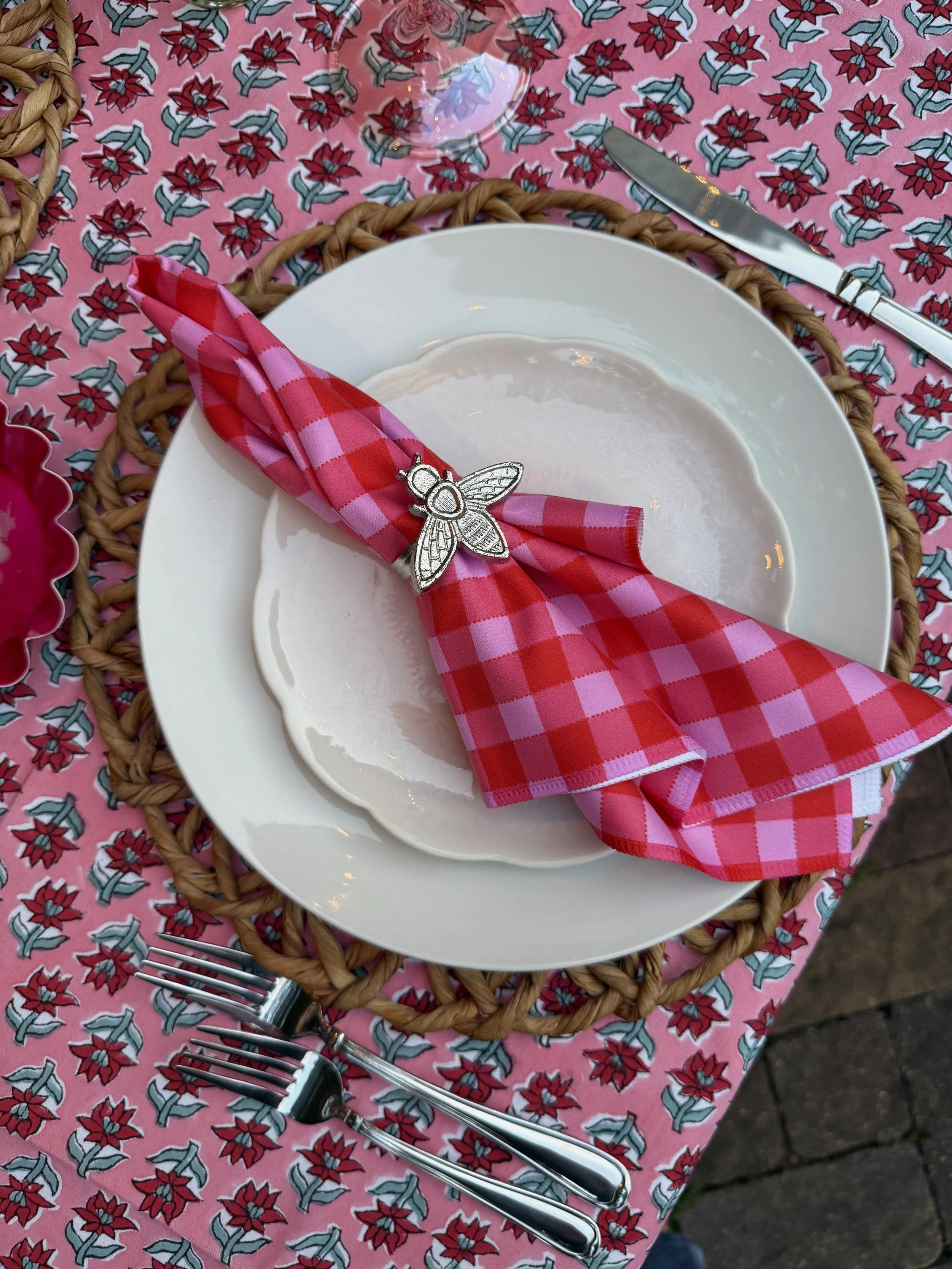 A table setting with a white plate, pink and red checkered napkin with a silver bee napkin ring, three forks to the left, a knife above, on a pink floral tablecloth, with a woven placemat underneath.