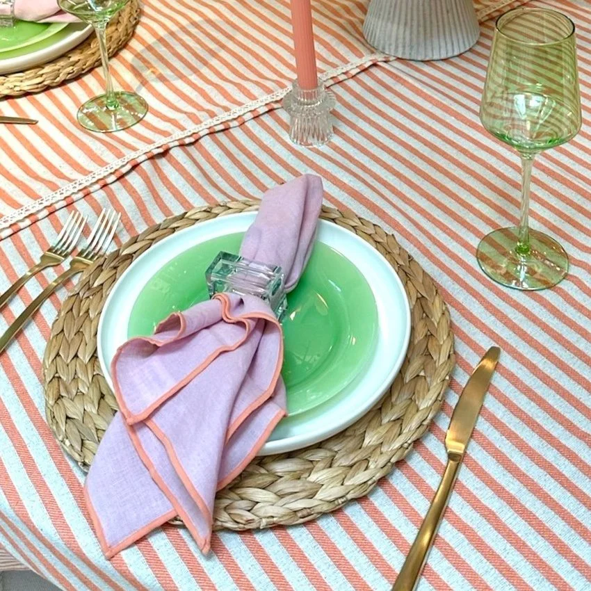 A table setting with a striped pink and white tablecloth, a woven placemat, a white and green plate, pink napkin, clear glassware, gold utensils, and a pink candle in a glass holder.