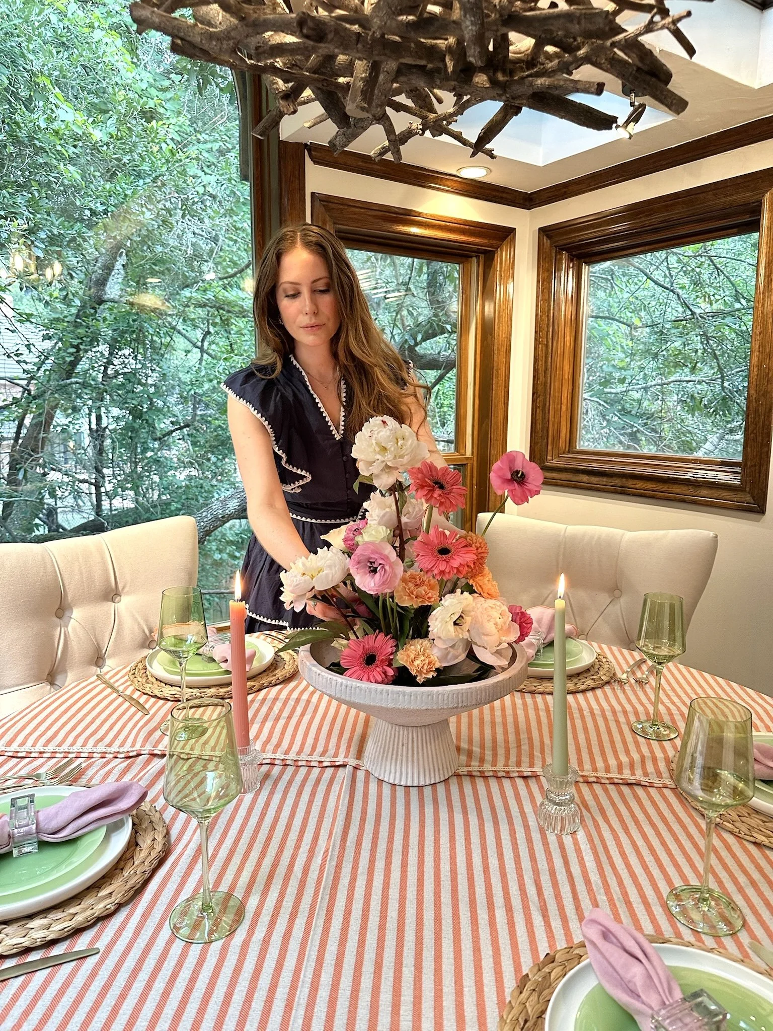 A woman with long brown hair arranging pink and white flowers in a large white vase on a dining table with a pink striped tablecloth. The table is set with green plates, pink napkins, wine glasses, and lit pink candles. The room has large windows showing green trees outside and a wooden chandelier hanging above.