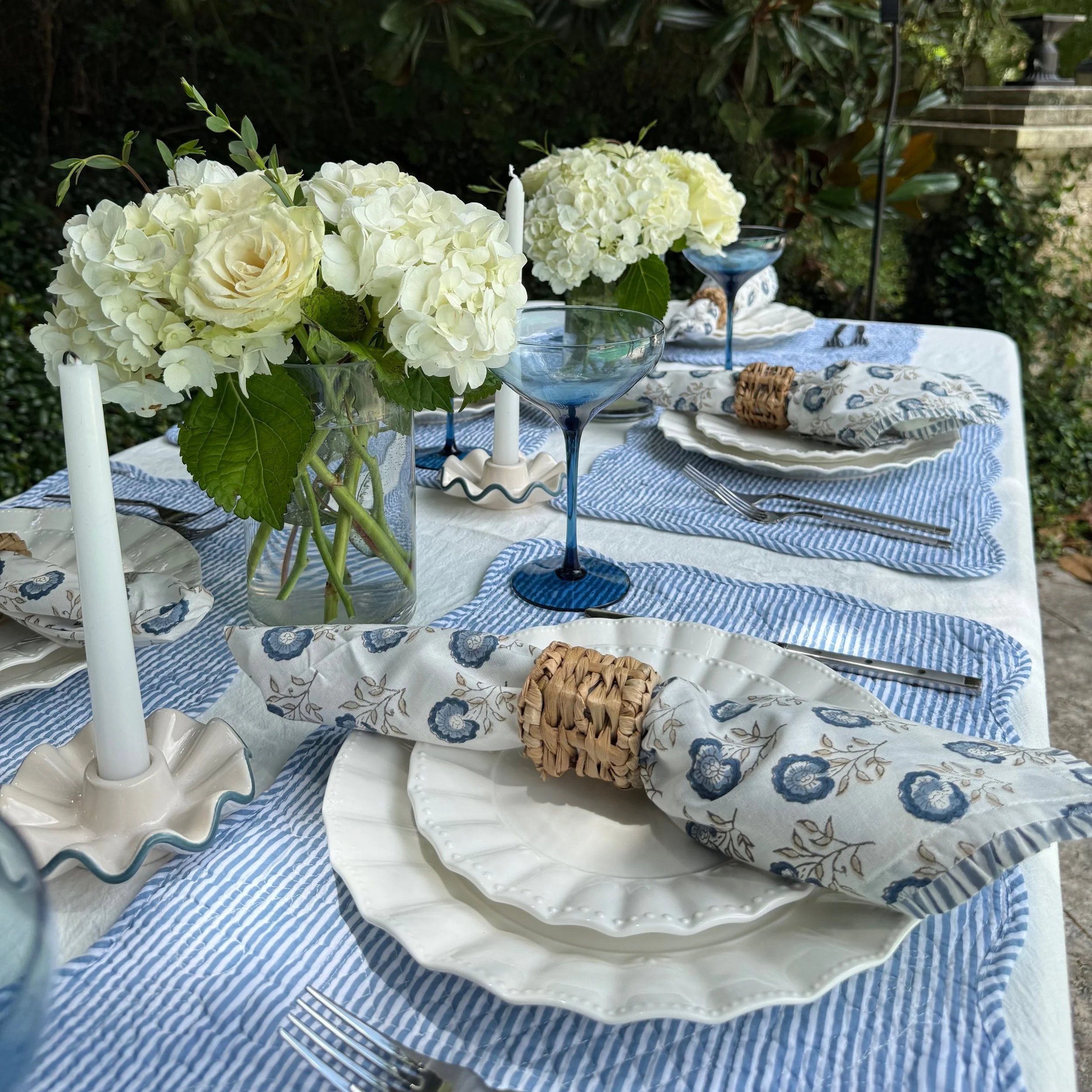 A table set with white and blue dinnerware, floral napkins, blue glassware, and floral centerpieces with white flowers, outdoors with greenery in the background.