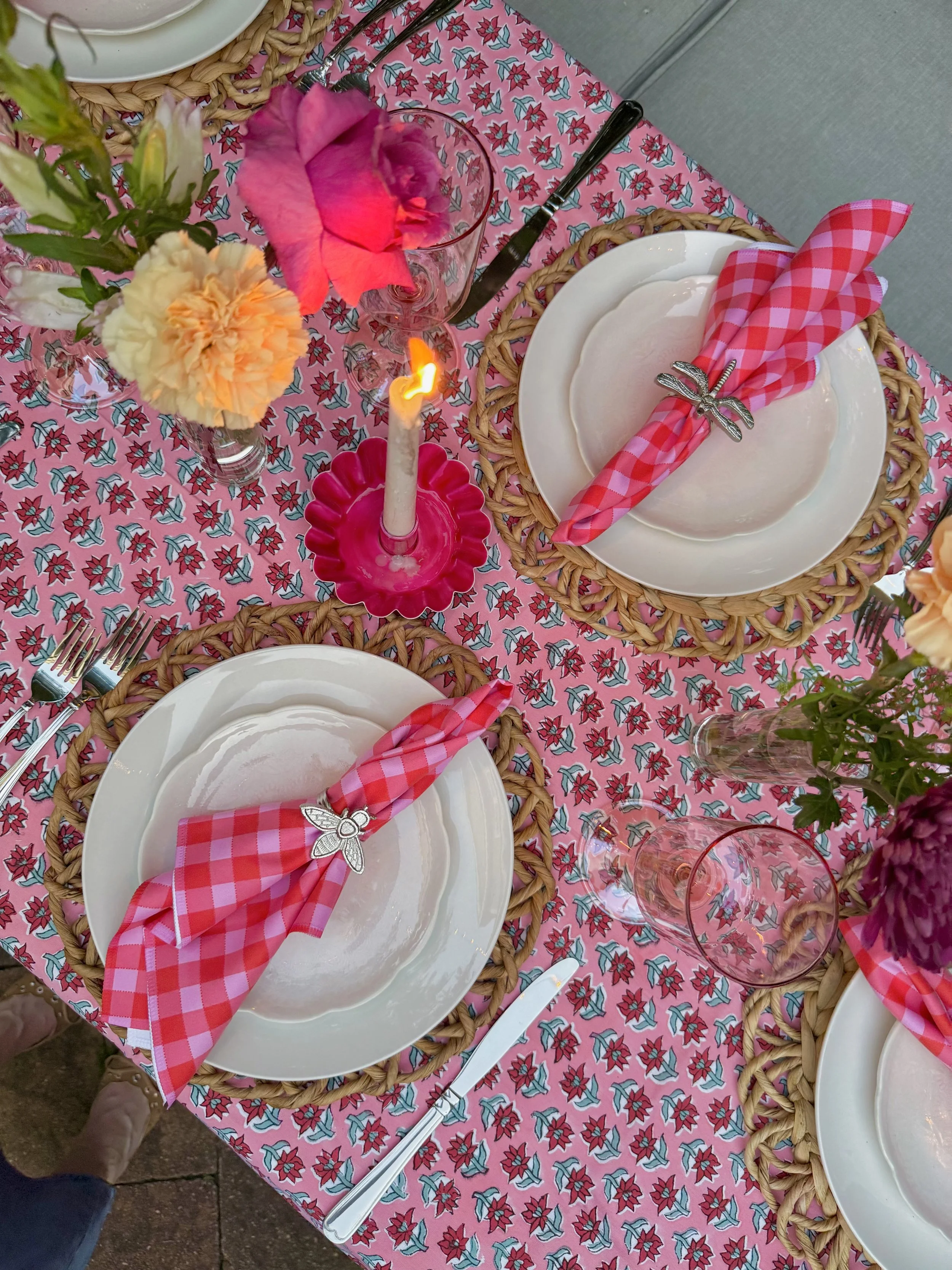 Table setting with floral tablecloth, white plates with pink and white checkered napkins held by silver rings with a bee design, clear glassware, pink lit candle in a pink dish, and flower vases with colorful flowers.