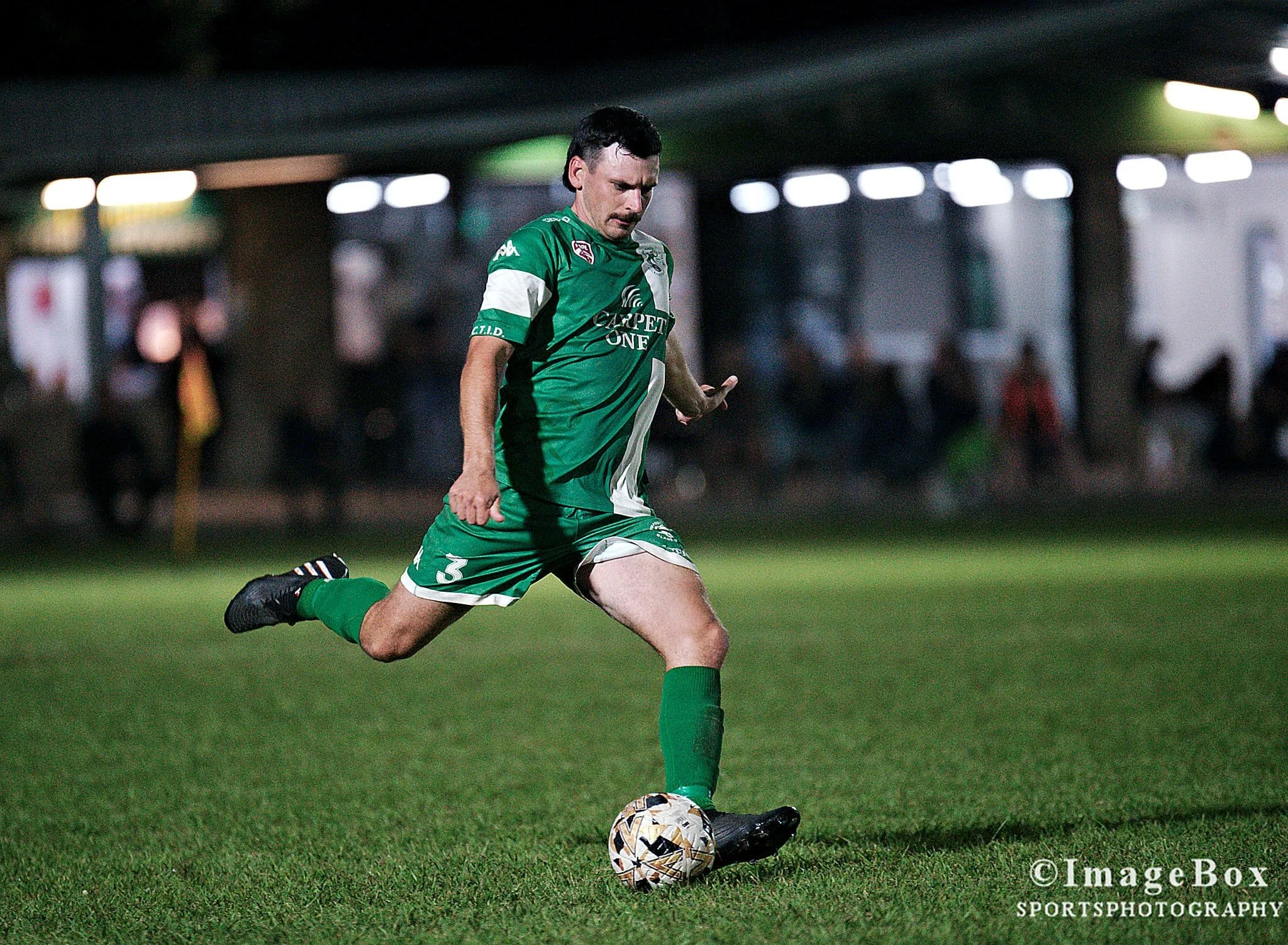 Soccer player in green uniform kicking a soccer ball on a grass field at night.