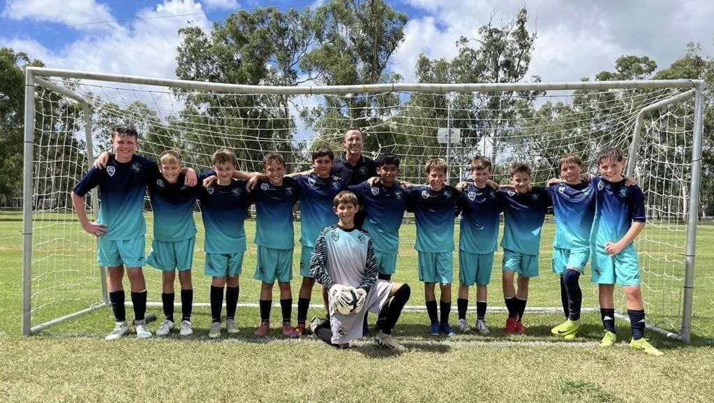 A youth soccer team of eleven boys and one coach, standing in a row inside a soccer goal on a grassy field, wearing matching blue uniforms, with a goalkeeper kneeling in front holding a soccer ball.