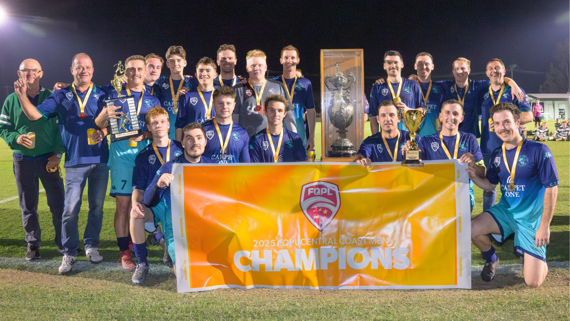 A group of soccer players and staff celebrating on a field at night, holding a large banner that reads '2025 FQPL Central Coast Men Champions,' with some players holding medals and trophies.
