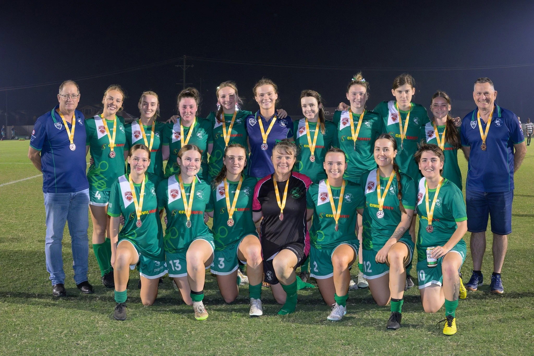 A women's soccer team posing on a field at night, wearing green jerseys, with some players kneeling in the front row and others standing, accompanied by two male coaches on either side, all wearing medals.