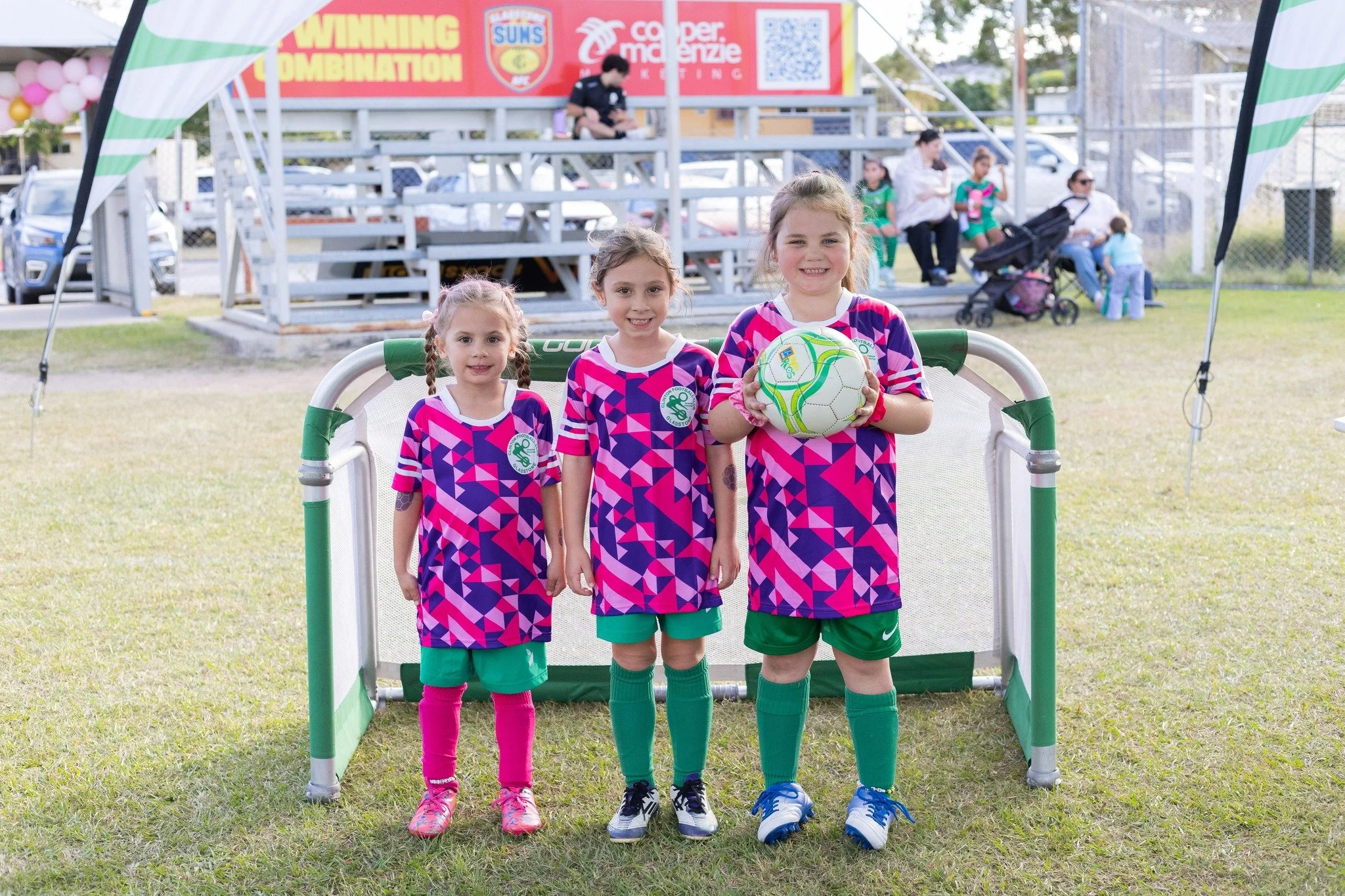 Three young girls in colorful soccer uniforms stand in front of a small goalpost on a grassy field, with one holding a soccer ball. In the background, there are bleachers with spectators, and banners advertising a sports brand and a club magazine.