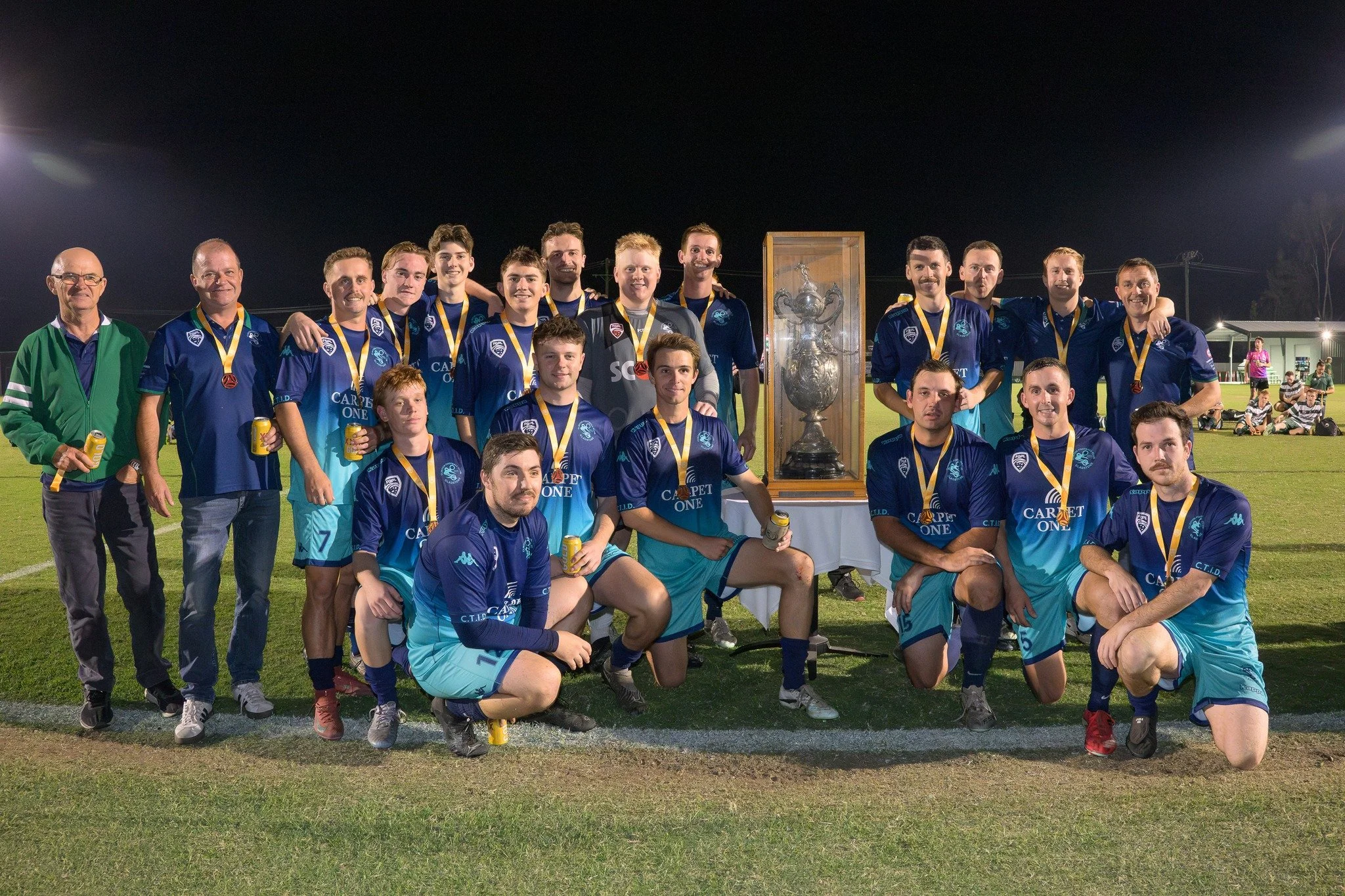 A soccer team with coaches posing on a field at night after winning a trophy, wearing blue and turquoise jerseys and medals.