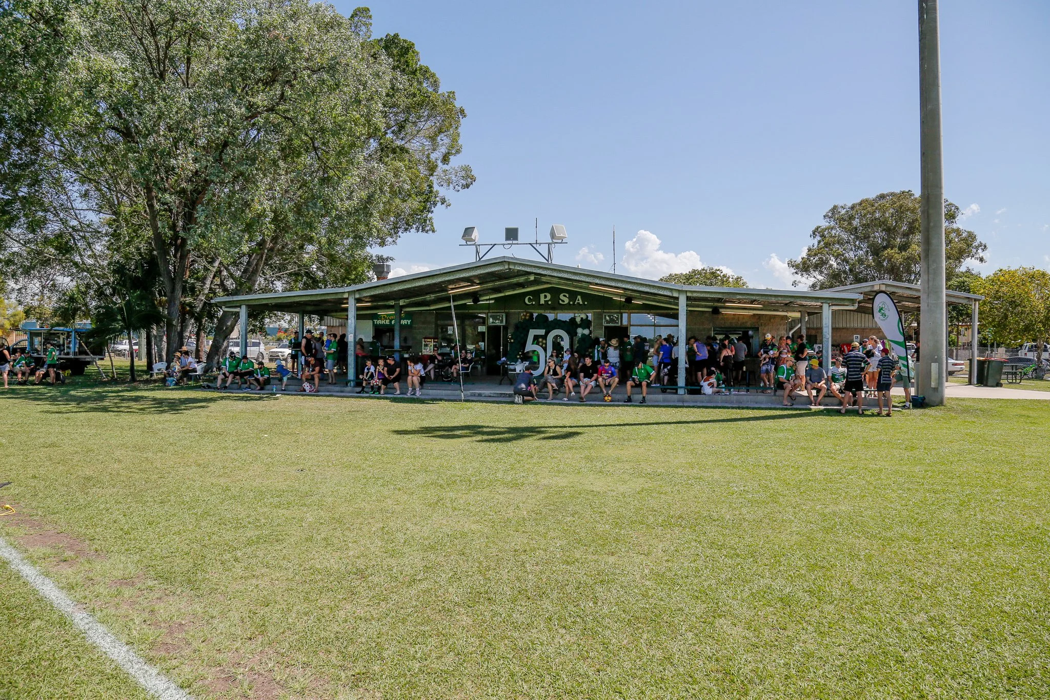 People sitting and standing outside a sports pavilion with 'C.P.S.A.' signage and a large '50' on the front, under a blue sky with trees and vehicles in the background.