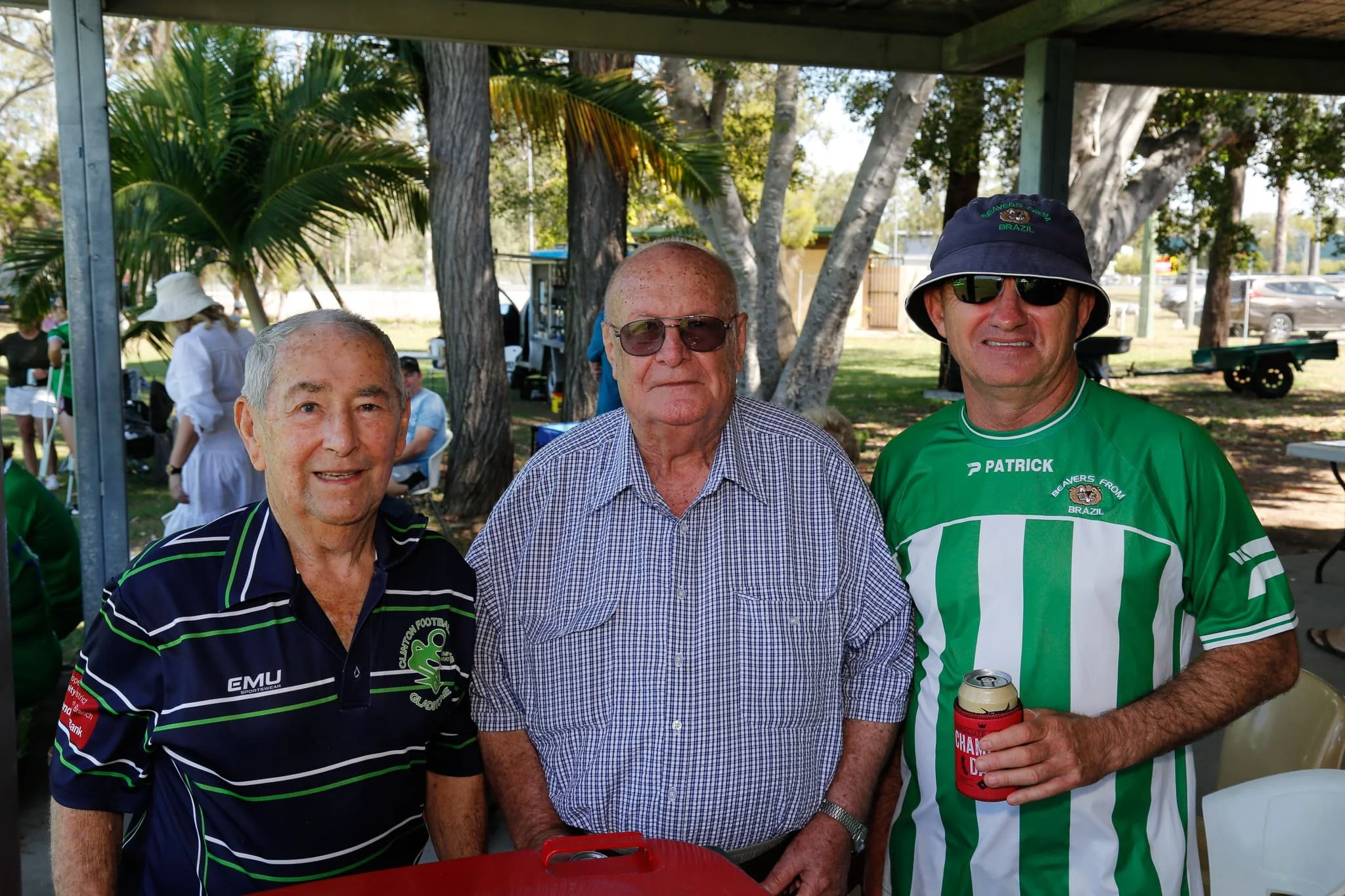 Three men standing outdoors at a gathering, with trees and other people in the background. The man on the left wears a striped sports shirt, the man in the middle wears a checkered shirt, and the man on the right wears a green and white striped sport