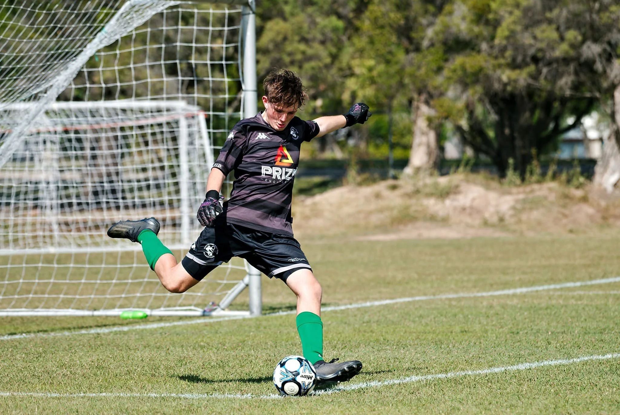 A soccer goalkeeper in black uniform kicks a soccer ball on a grass field near the goalpost.