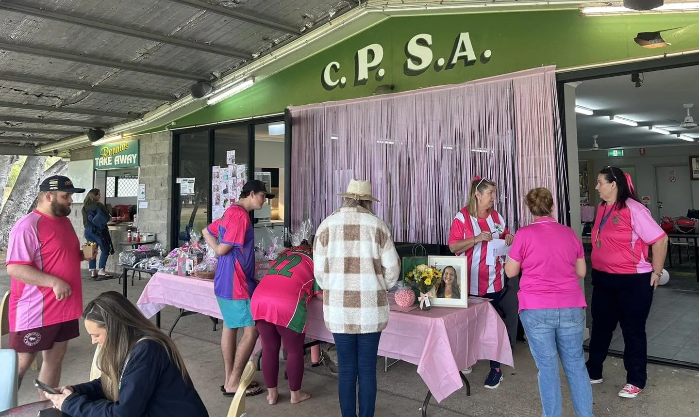 People gathered outside a building with a green sign reading 'C.P.S.A.' and a smaller sign for 'Donuts Take Away.' They are wearing pink shirts, some are talking at a table covered with a pink cloth, and there are items like a framed photo, flowers, 