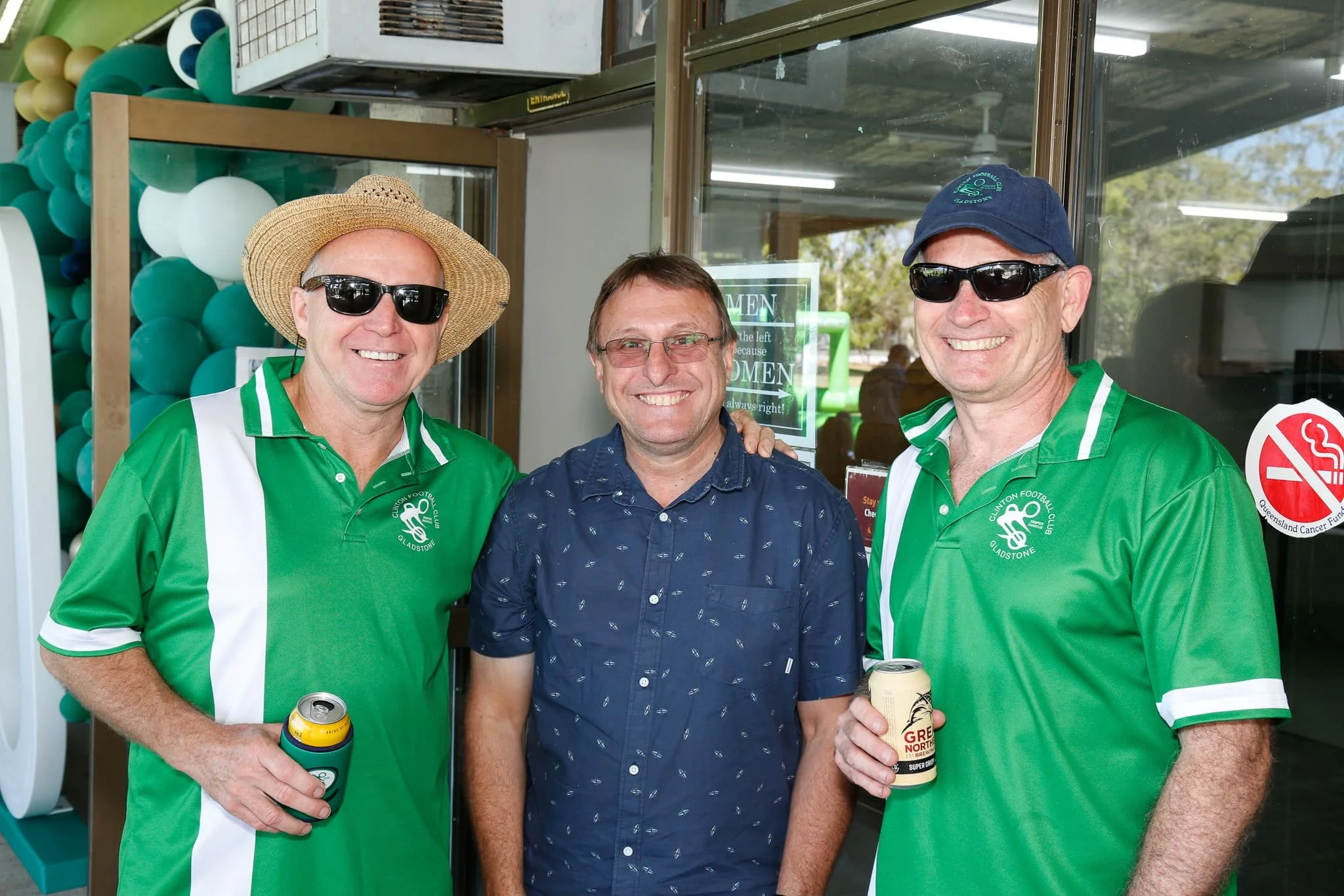 Three middle-aged men standing together smiling, two wearing green sports jerseys with QLD Gladstone logos, sunglasses, and holding cans of beer, one in a blue patterned shirt, in a casual indoor setting with balloons and a sign on the door in the ba