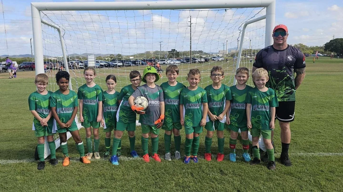 Children in green soccer uniforms standing in a row on a soccer field with a coach on the right, behind a goal, on a sunny day.
