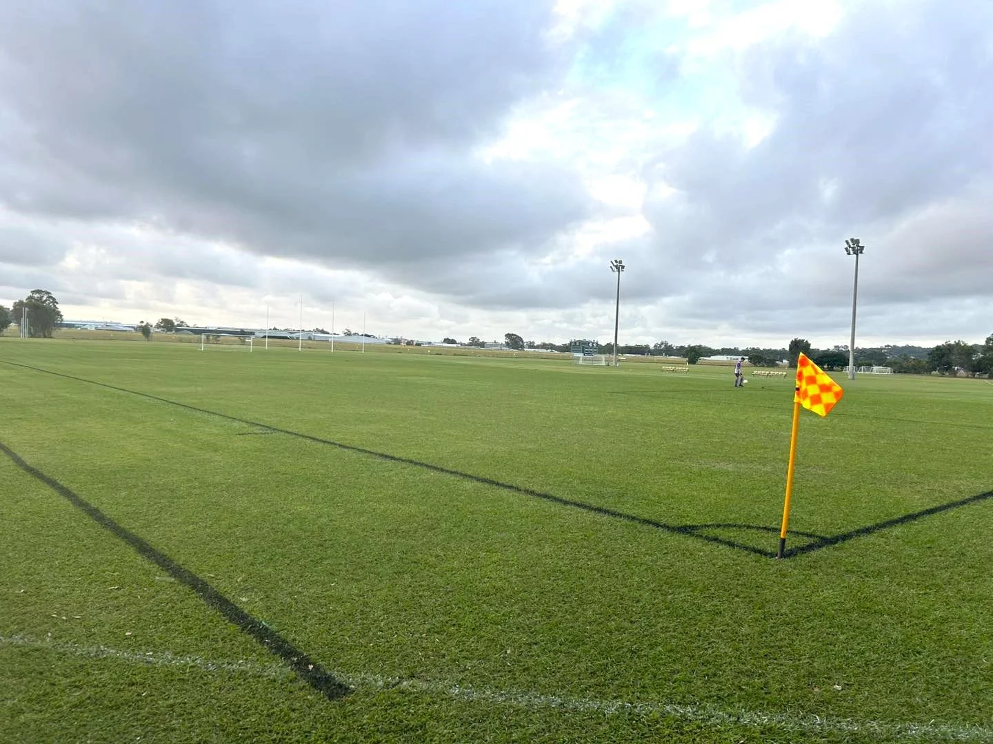 A sports field with green grass and marked lines, surrounded by tall light poles, under a cloudy sky, with a corner flag in the foreground.