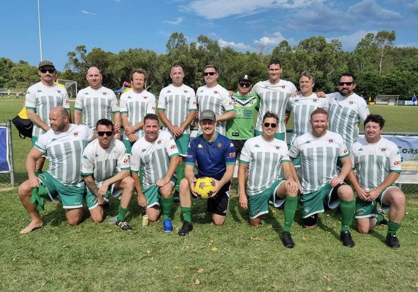 Group of men in soccer uniforms posing on a grassy field with trees and blue sky in the background. Some are standing, some kneeling, with a soccer ball held by a man in the center wearing a blue jersey.