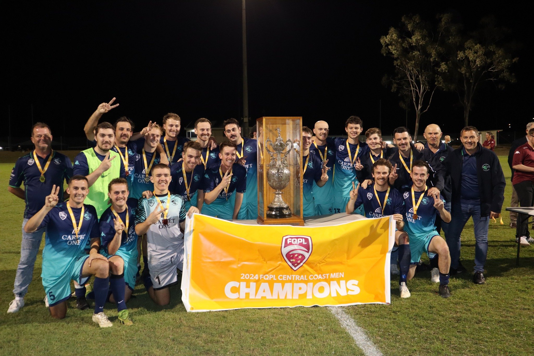 Soccer team celebrating as 2024 FQPL Central Coast Men Champions, group photo on field at night with trophy, team members wearing medals, holding a shield-shaped banner, with two coaches or staff members, behind a wooden display case with a trophy.