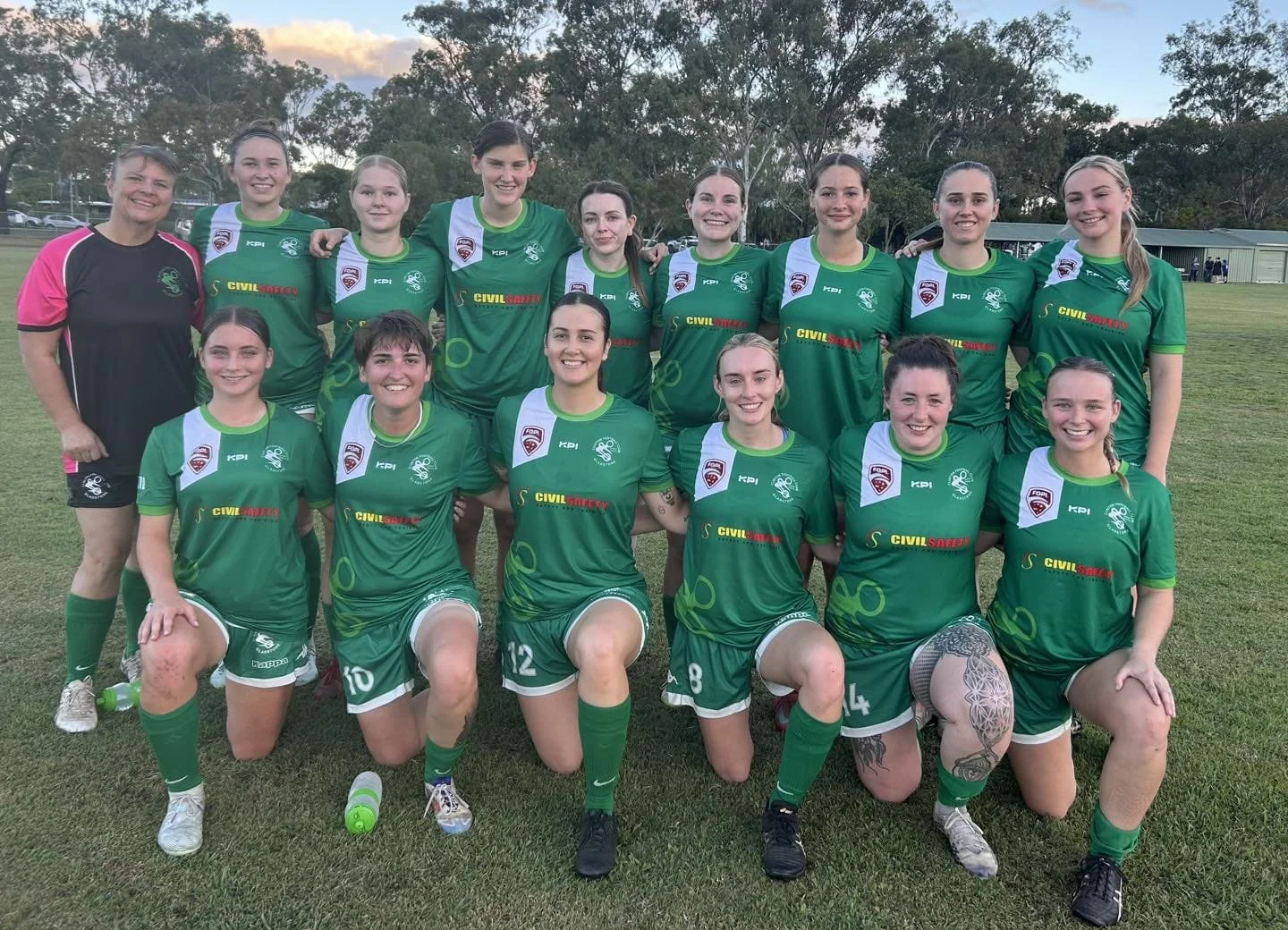 A women's soccer team poses on a field, wearing green uniforms with white accents, along with their coach or team official on the side, all smiling after a game or practice.
