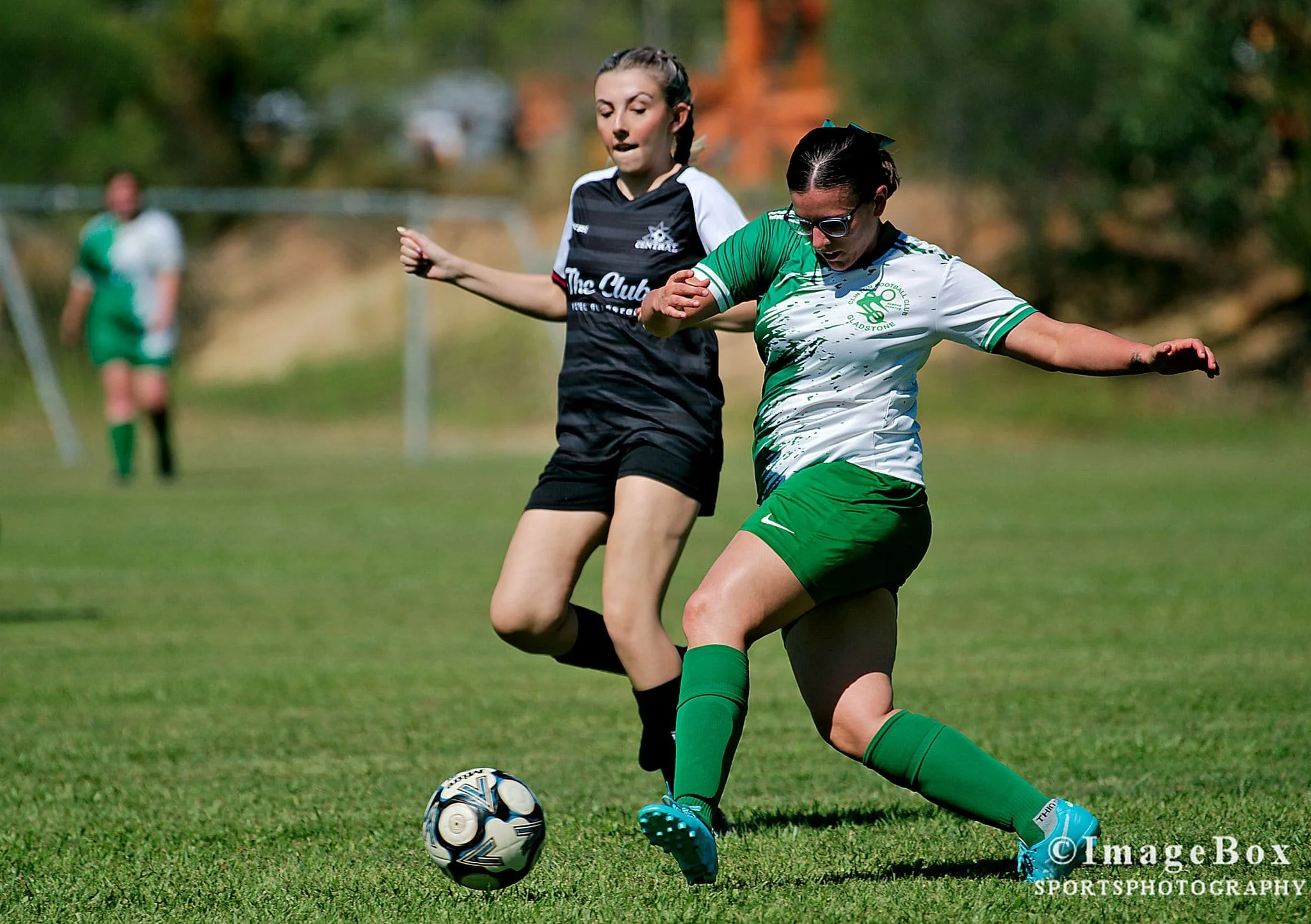 Two female soccer players competing for the ball during a match on a grassy field with a blurred opponent and goalpost in the background.