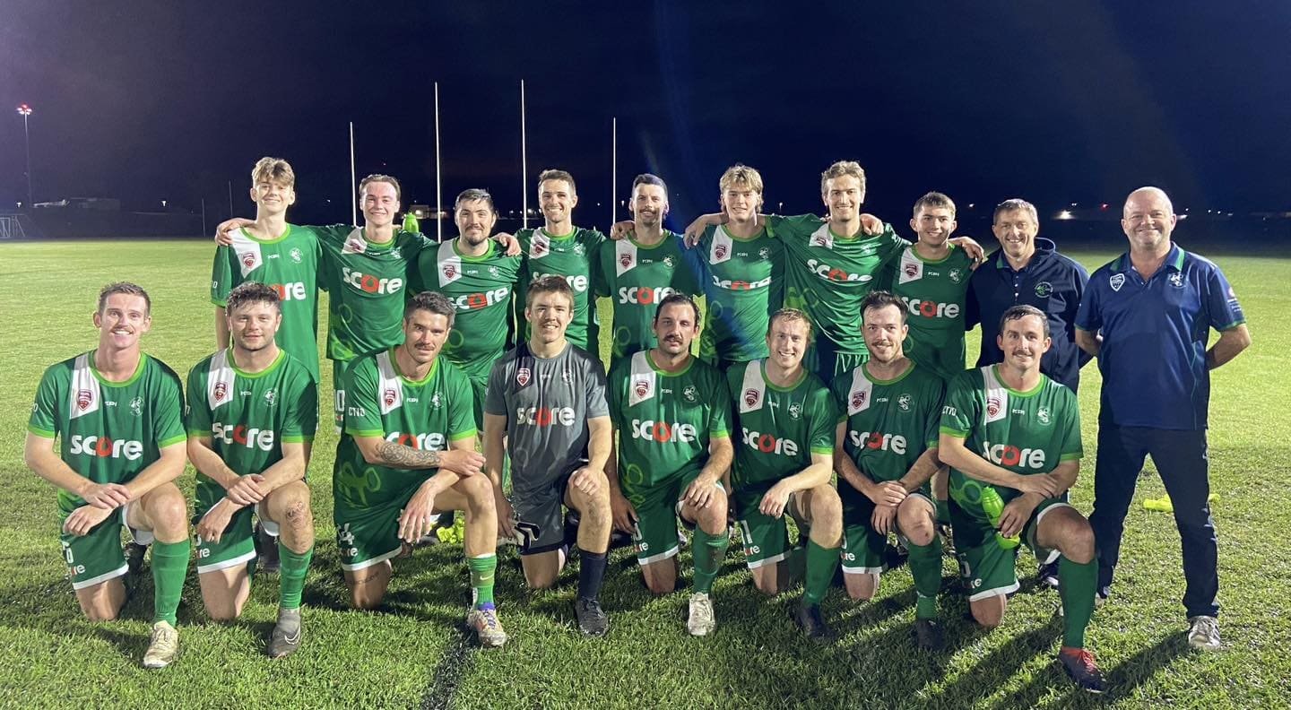 A rugby team in green uniforms poses for a photo on a field at night, with coaching staff in blue jackets, in front of rugby goalposts.
