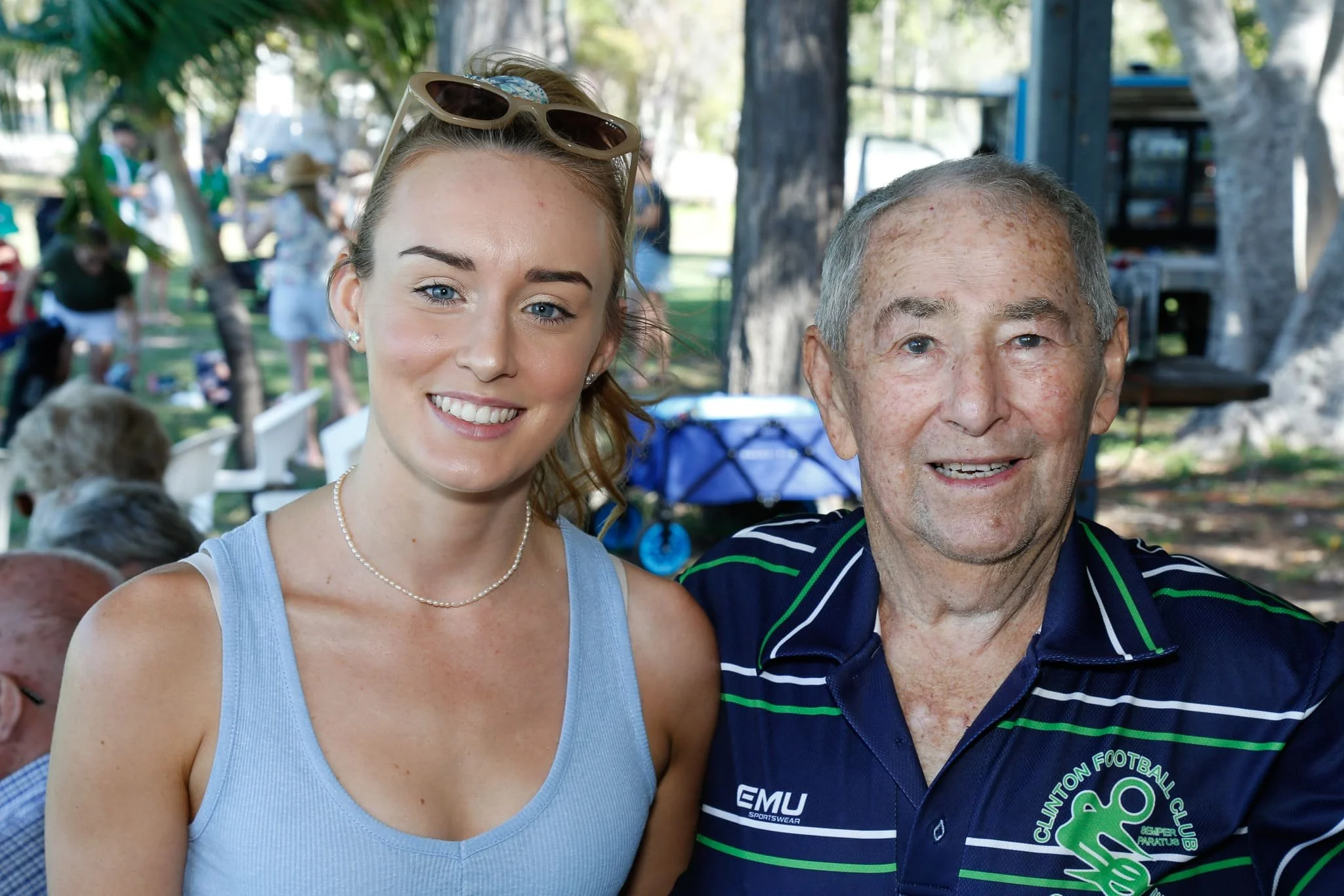 A smiling young woman with sunglasses on her head and a pearl necklace, and an older man in a navy and green Clontarf Football Club jersey, pose together outdoors at a park or picnic area with trees and other people in the background.