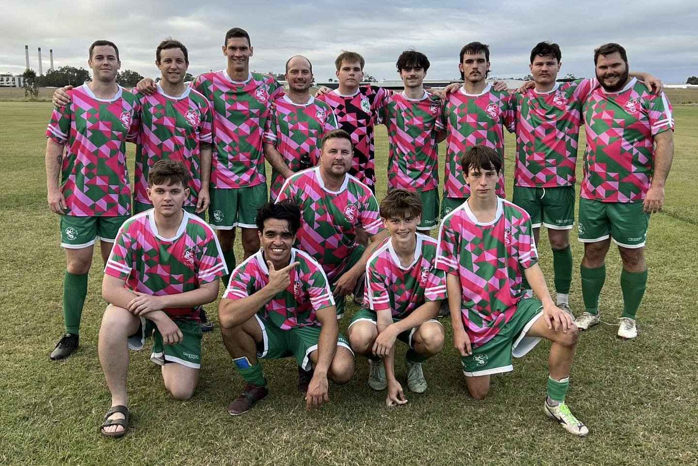 A soccer team of young men and boys in colorful pink, green, and black jerseys, posing for a team photo on a grassy field with a cloudy sky. They are arranged in two rows, some standing and some kneeling.