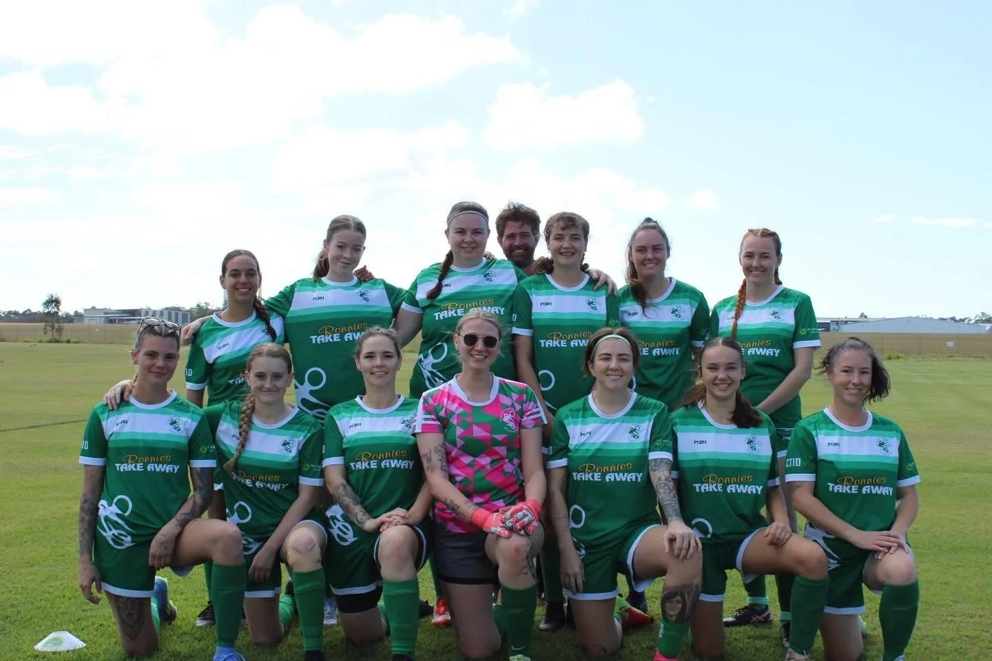 A women's rugby team in green jerseys with white stripes, posing on a grassy field with a blue sky and clouds in the background.