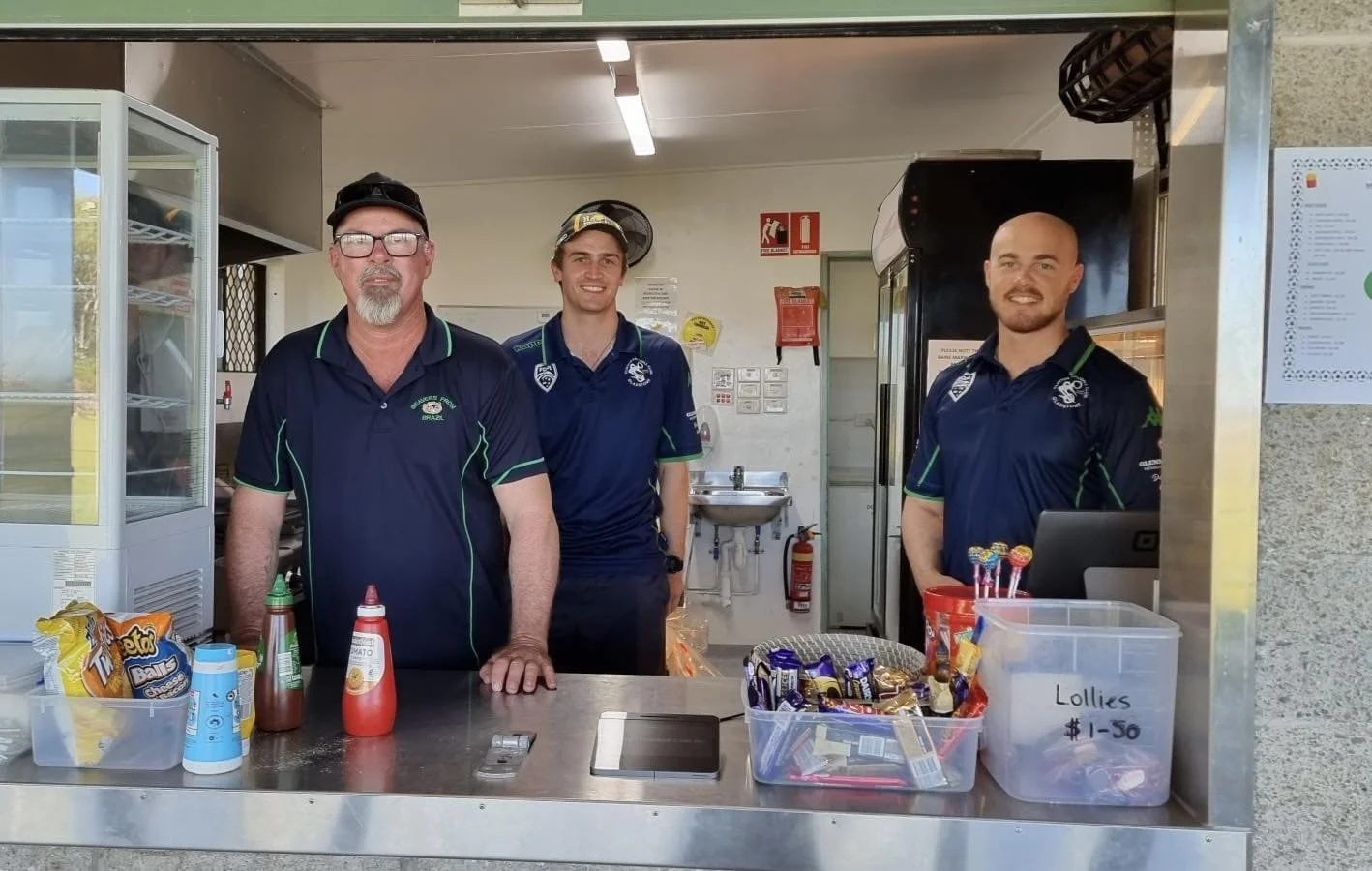 Three men in navy blue uniforms stand behind a snack counter with various snacks and condiments.