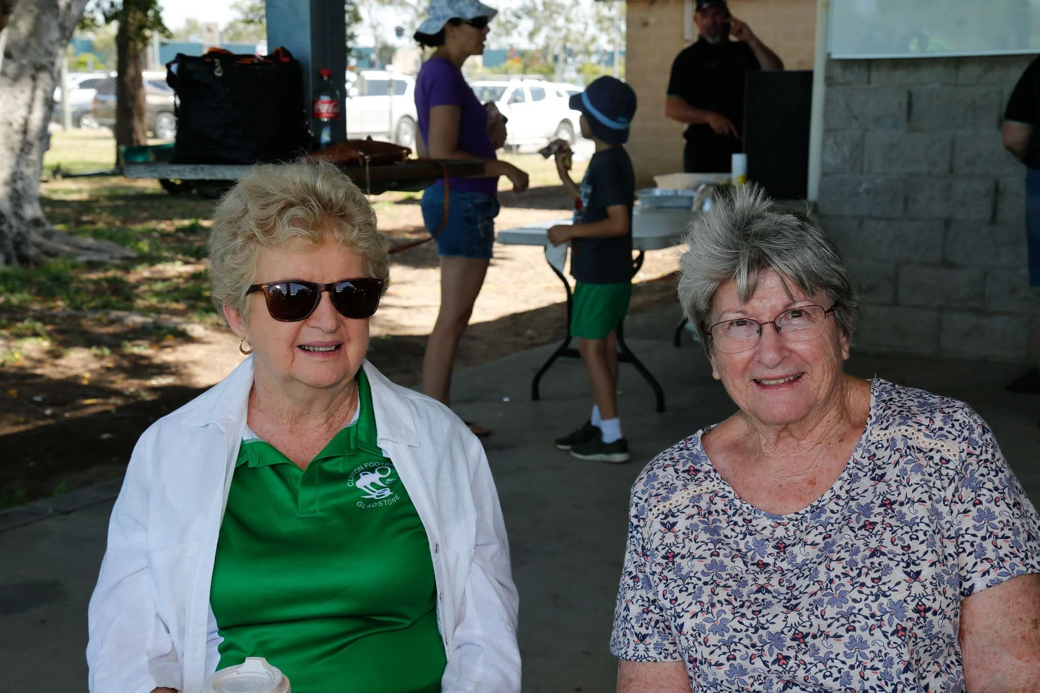 Two elderly women sitting outdoors at a social gathering, with one wearing sunglasses and a green shirt, and the other wearing glasses and a patterned top, smiling at the camera.