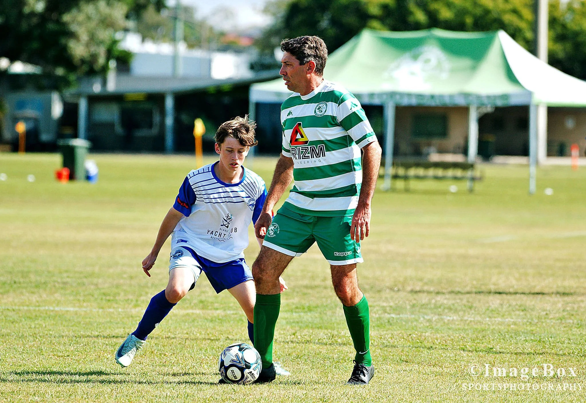 A man and boy playing soccer on a grassy field. The man wears a green and white striped jersey with green shorts, while the boy wears a white and blue jersey with white shorts. They are competing for the ball.