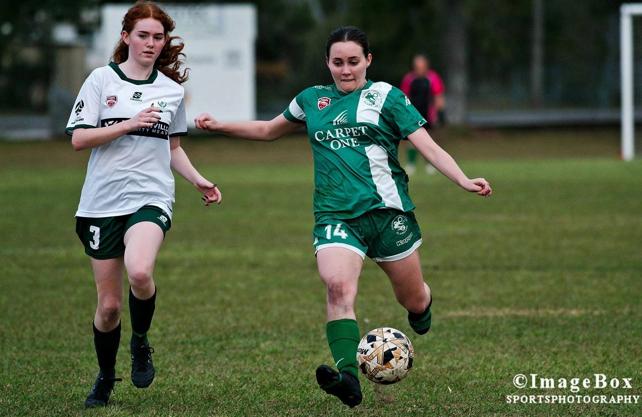 Two female soccer players competing for the ball on the field, with a background of a goalpost and trees.