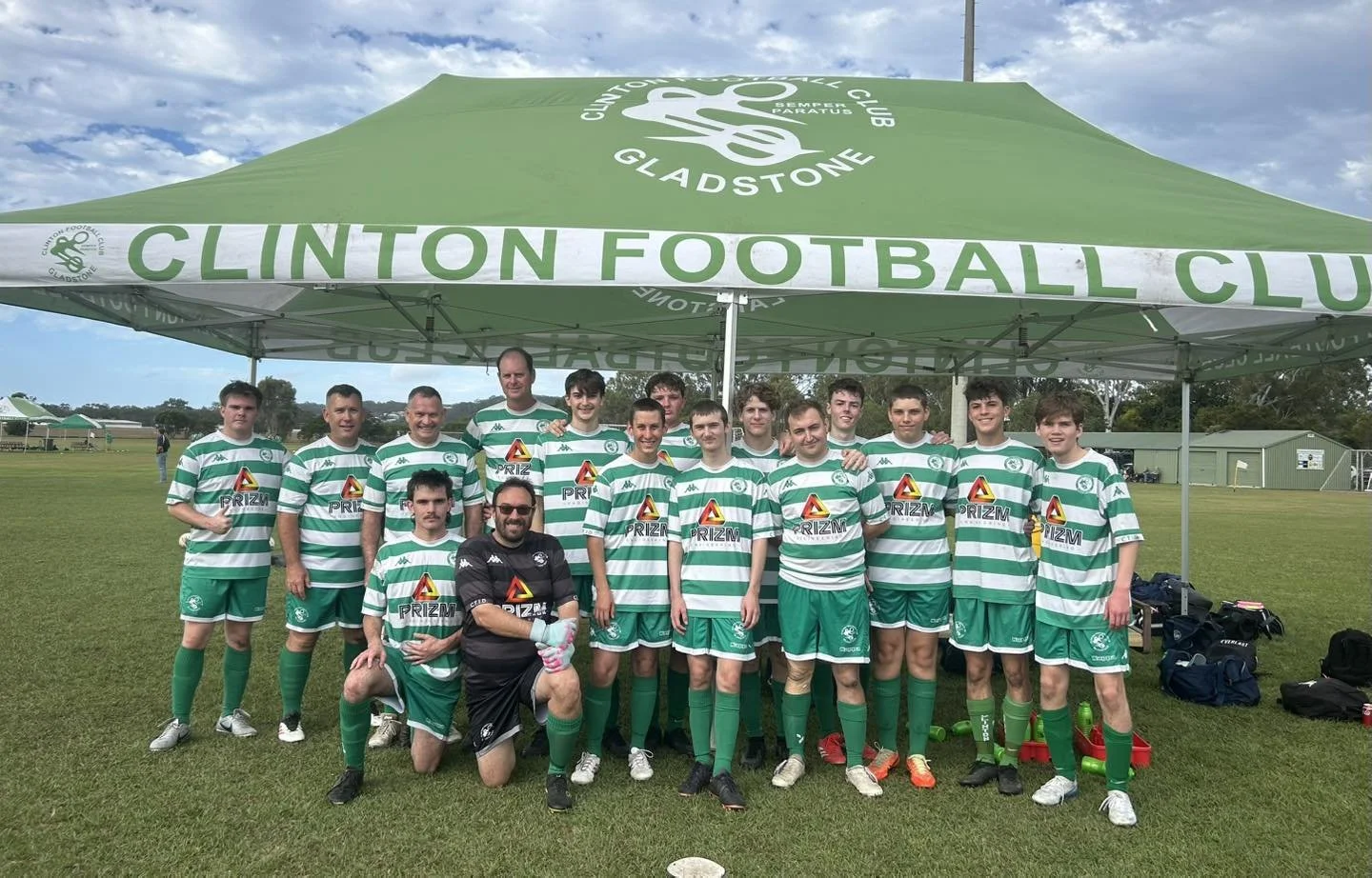 A group of young football players and their coach standing under a large green tent that reads 'Clinton Football Club' in white, on a grassy field, with some bags and water bottles nearby.