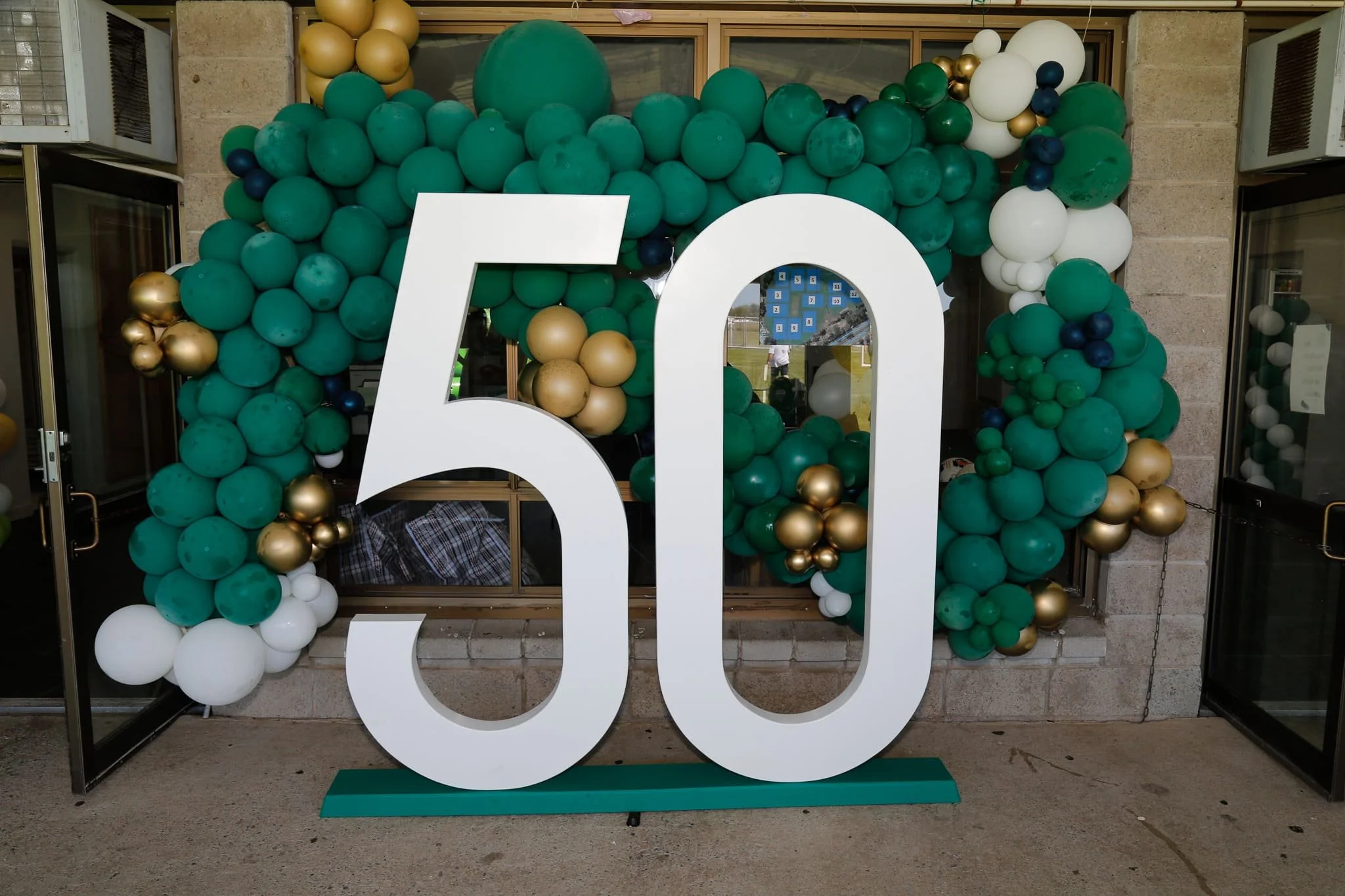 Large white number '50' with a balloon arch consisting of green, white, gold, and navy balloons behind it, likely celebrating a 50th anniversary or birthday event.