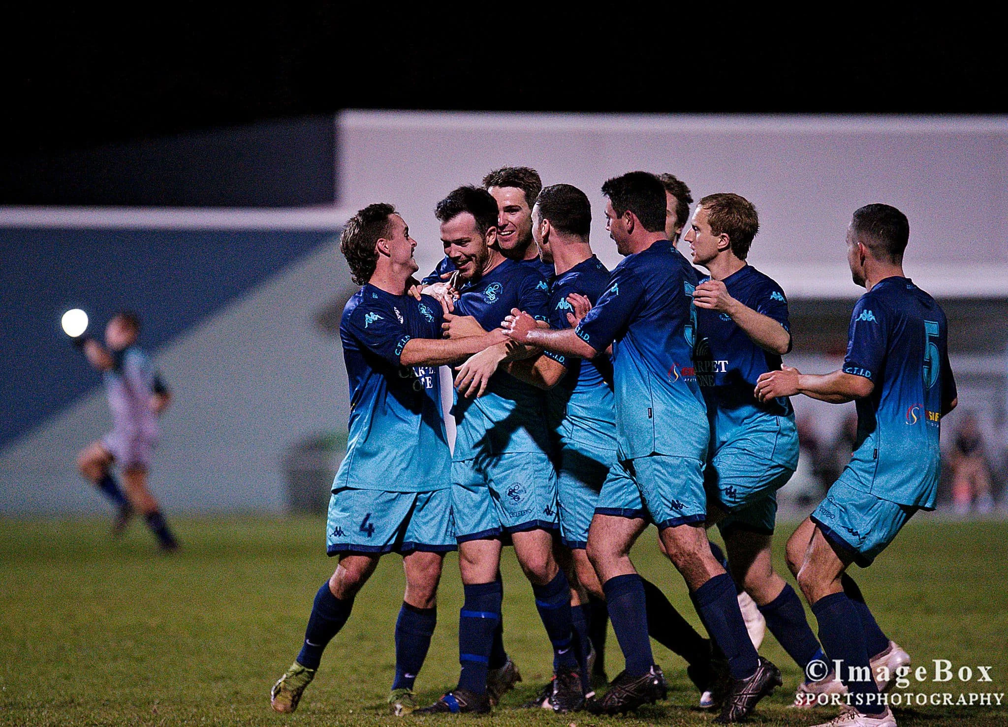 Group of soccer players wearing blue uniforms celebrating together on the field at night.