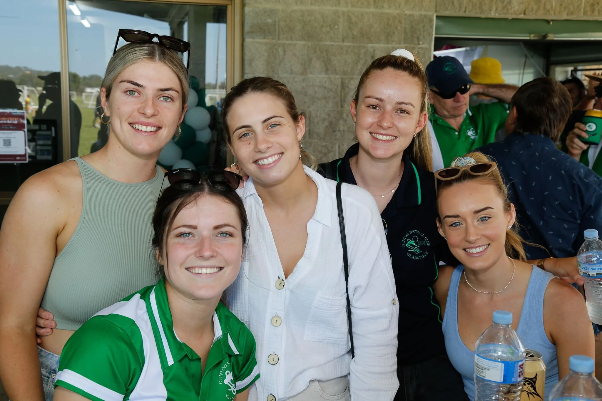 Group of six young women smiling at an outdoor event, some wearing casual clothes and sunglasses, with drinks and people in the background.