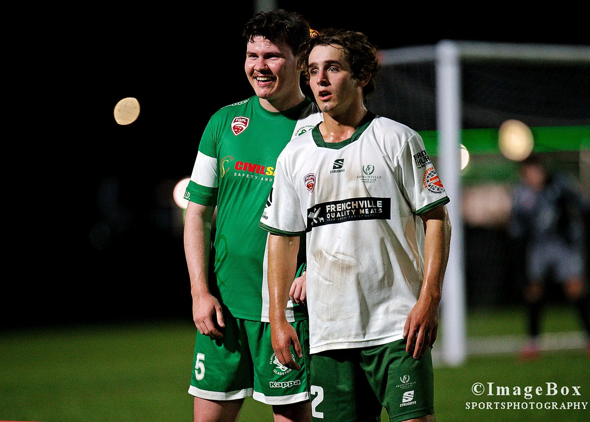 Two young male soccer players stand on a field at night, one in a green uniform smiling, the other in a white uniform looking serious, with a goalpost and blurred spectators in the background.