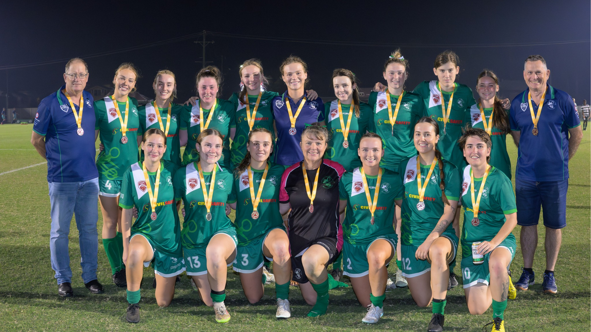 A women's soccer team celebrating after a game, wearing green jerseys with medals around their necks, posing on a soccer field at night with coaches.