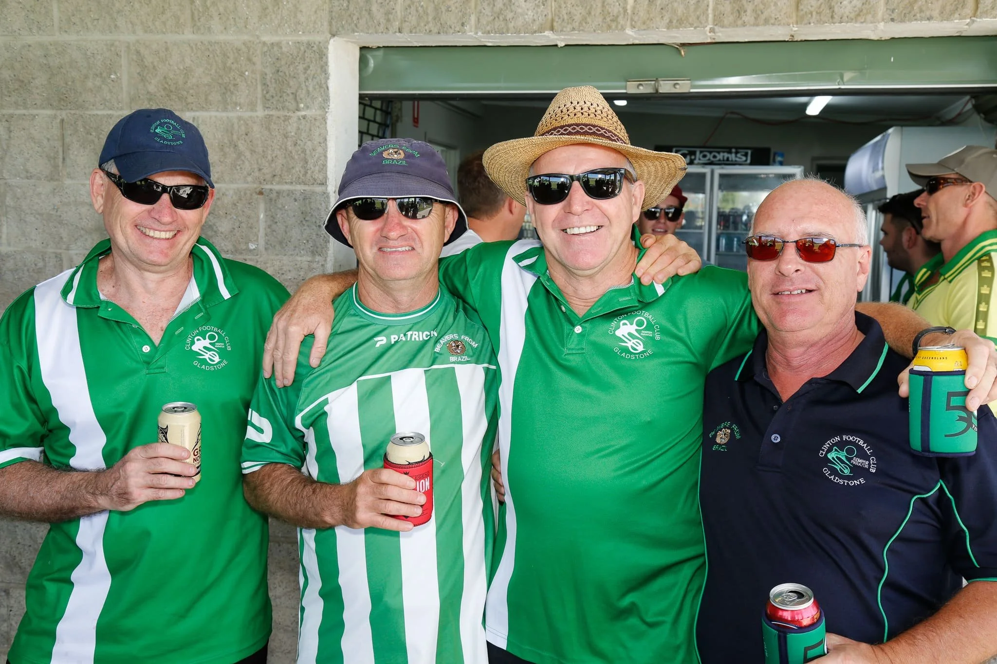 Four men standing together, smiling, wearing Gladstone Football Club jerseys with drinks in hand, posing indoors with a sports bar or club setting in the background.