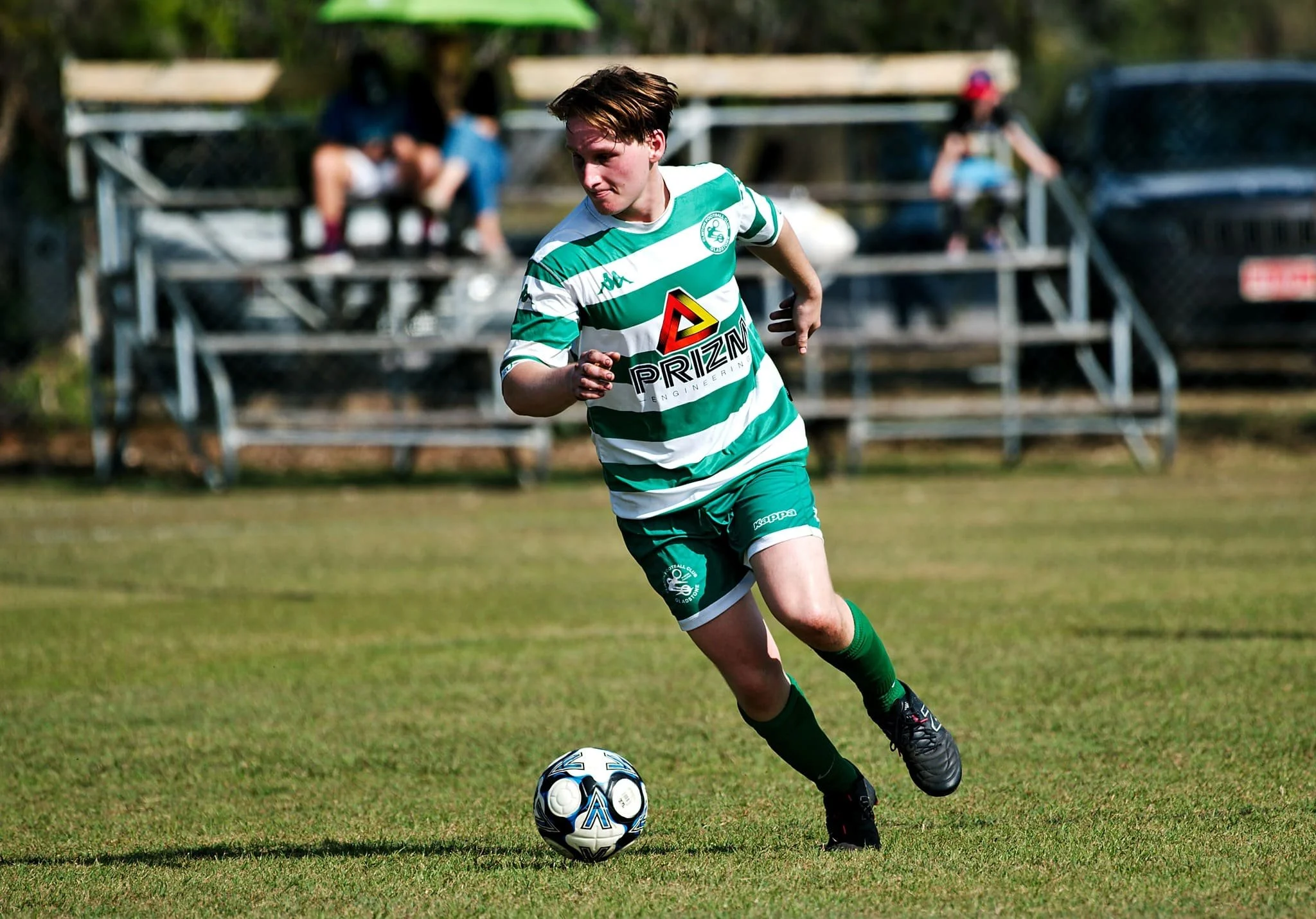 A young male soccer player in green and white striped uniform running with a soccer ball on a grassy field.