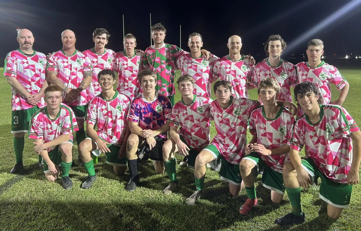 Group of young male soccer players in pink, white, and green jerseys, some kneeling, some standing, on a grassy field at night, with stadium lights in the background.