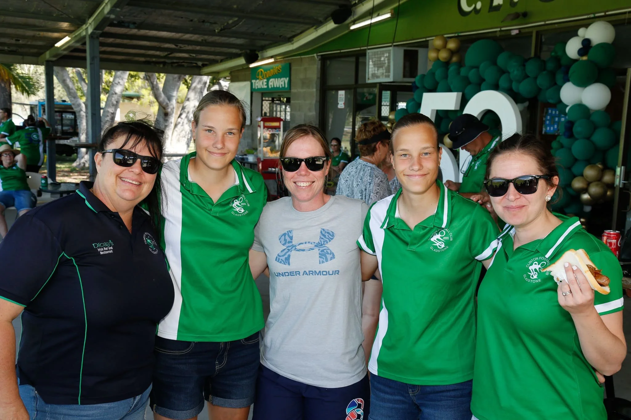 Group of five women, four wearing green sports jerseys, standing together at an outdoor event, smiling, with balloons and a sign in the background.